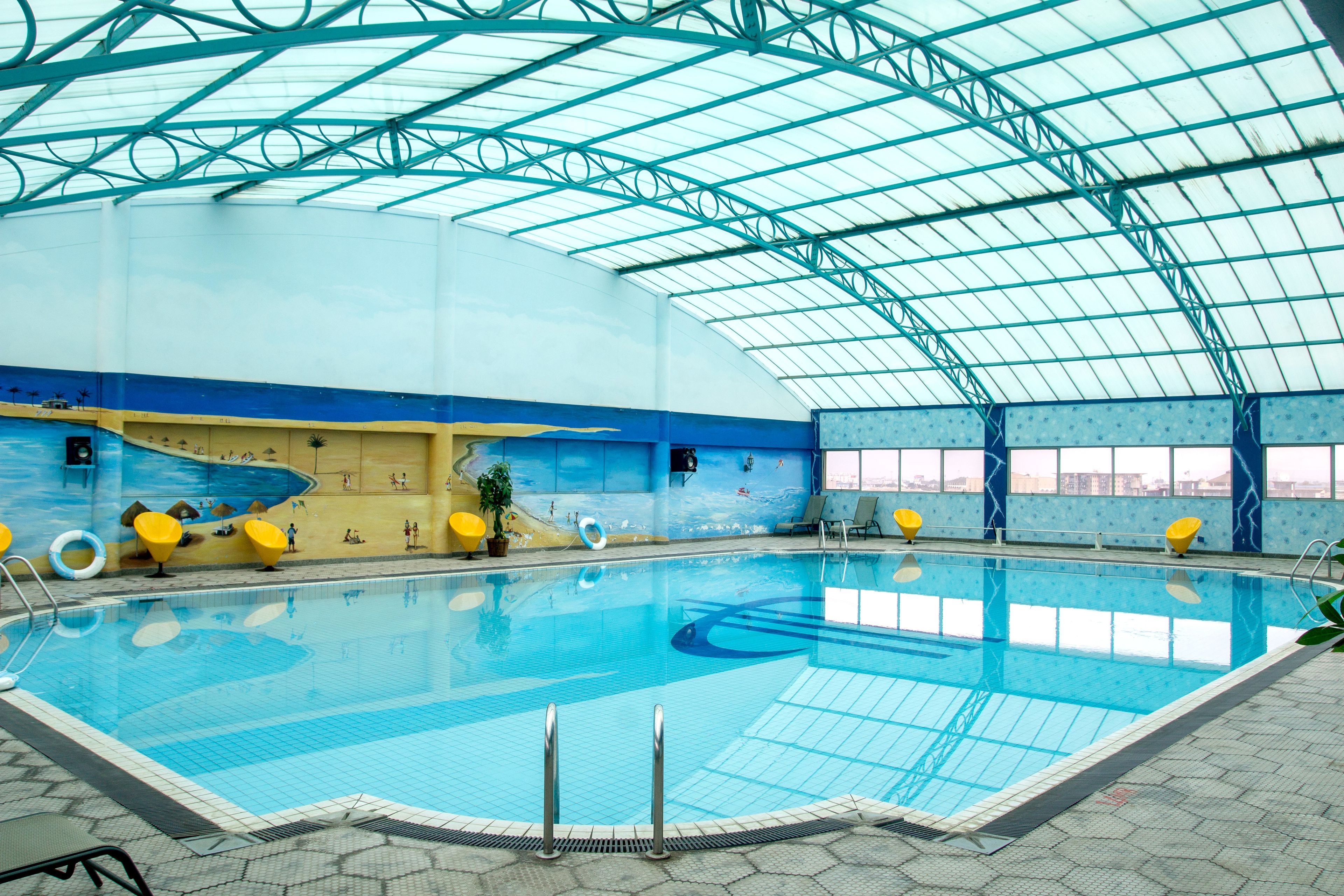An indoor swimming pool with a high ceiling and clear glass panels, surrounded by yellow chairs and a mural on the walls.
