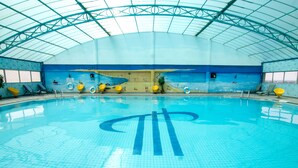 En indendørs swimmingpool med et stort muralmaleri, krystalklart blåt vand og solformede dekorationer.