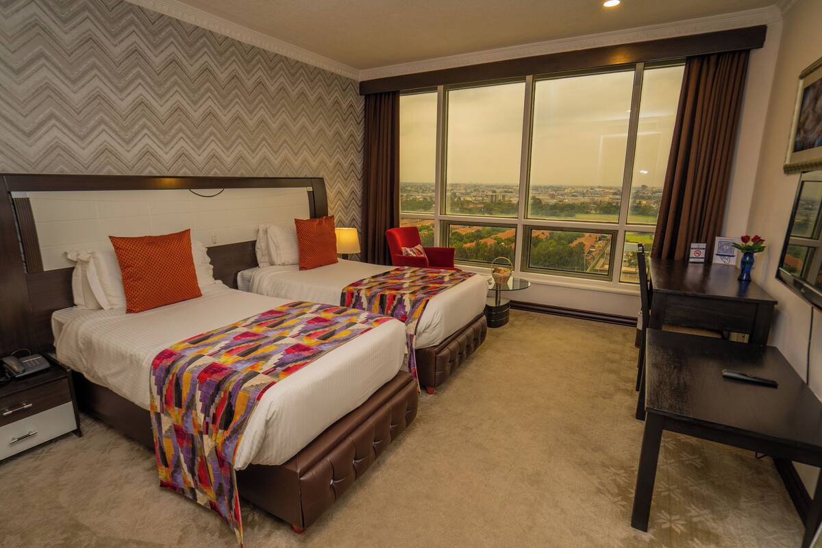 A hotel room with two beds, a desk, a chair, and a large window offering a city view.