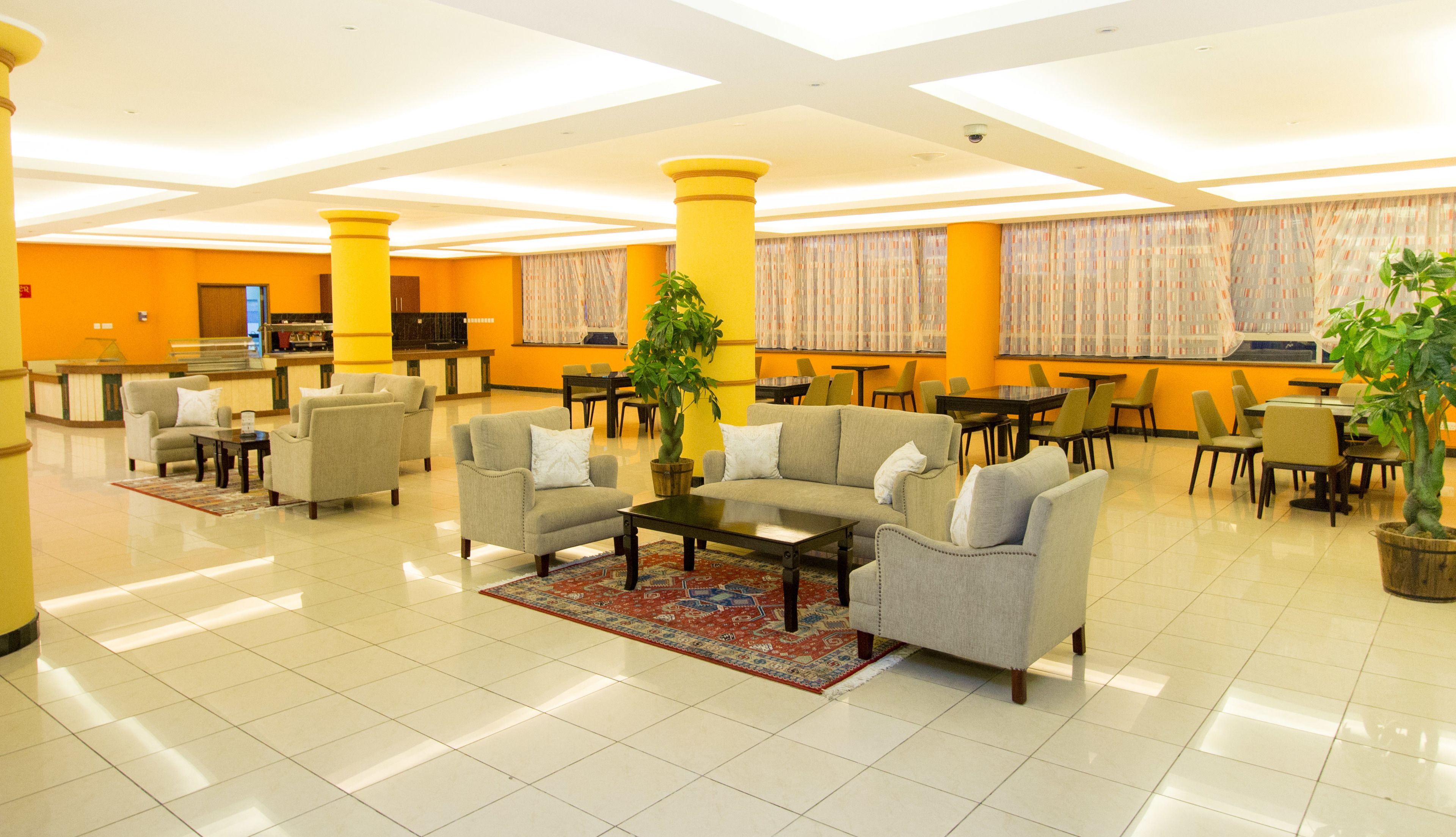 A spacious lobby with multiple seating areas, including sofas and chairs, arranged around a central coffee table. There are large windows with curtains, a reception desk, and a plant in the center.