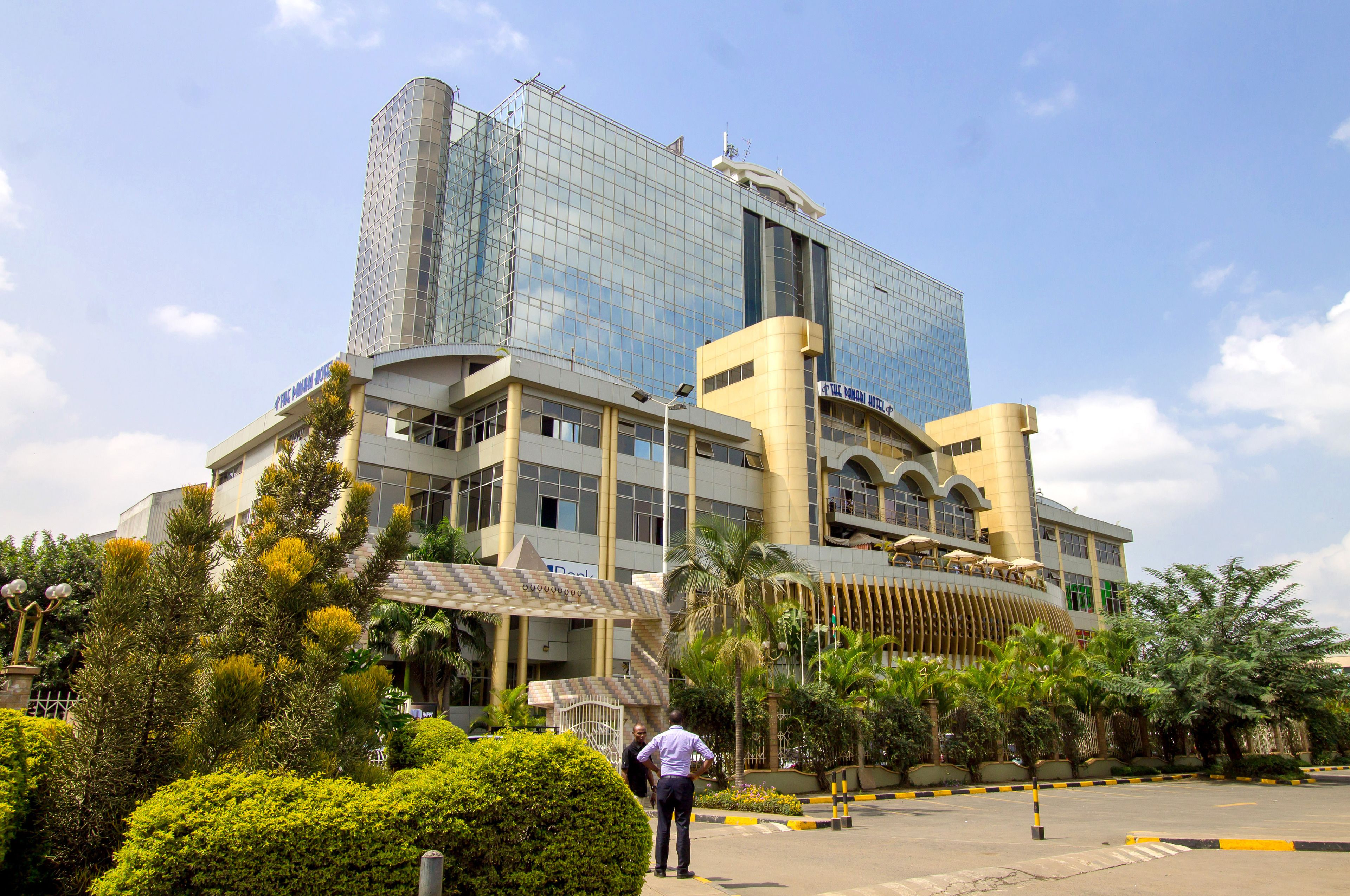 A modern hotel with a glass facade, a central courtyard with palm trees, and a parking area.