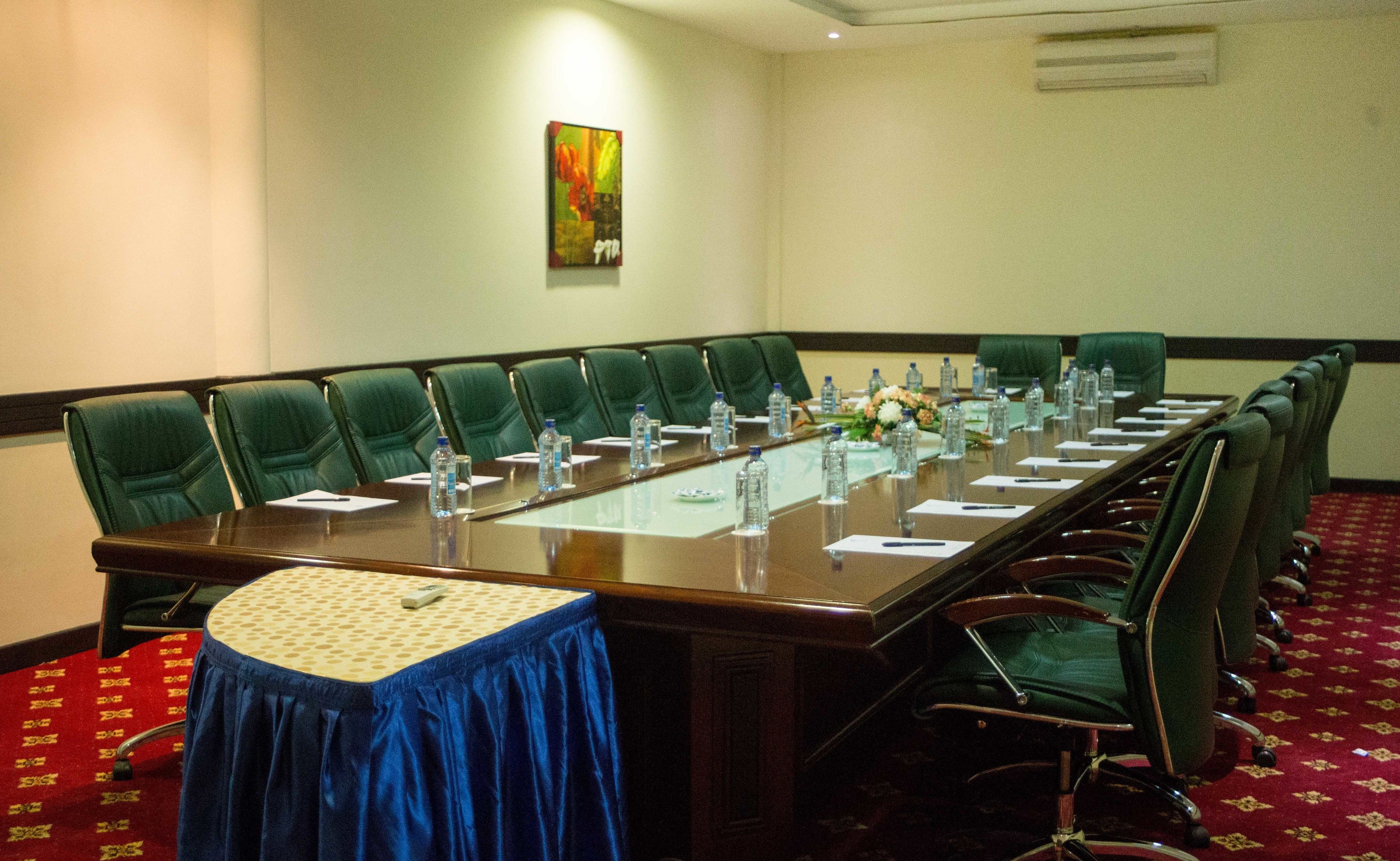 Un salón de conferencias con una larga mesa, sillas verdes, botellas de agua y un mantel azul.