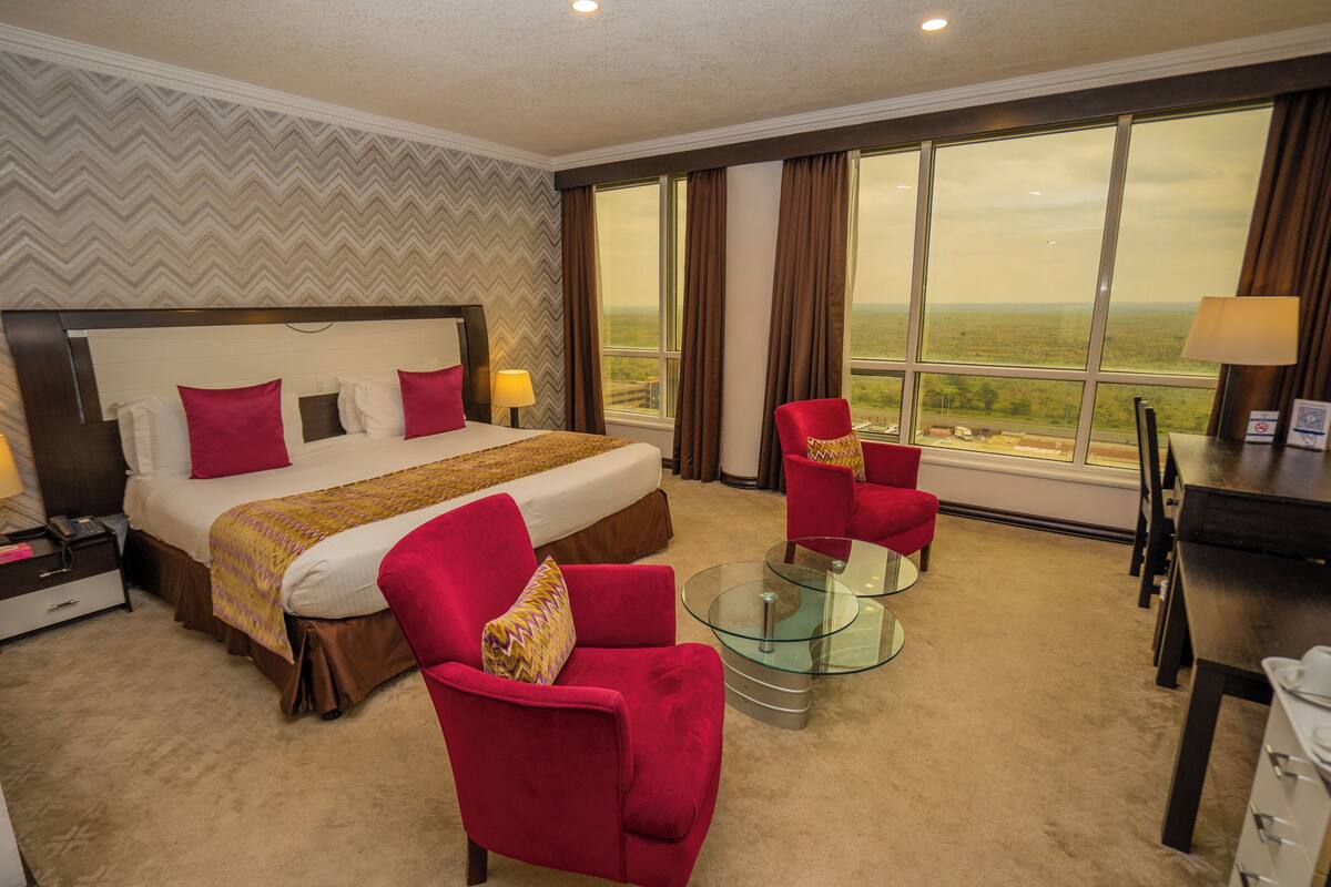 A hotel room with a large bed, two red armchairs, a glass coffee table, and a view of the countryside.