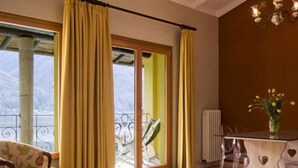 A room with a large window, yellow curtains, a chair, a table with a vase of flowers, and a radiator. - Residenza Lago di Lugano (Porlezza)