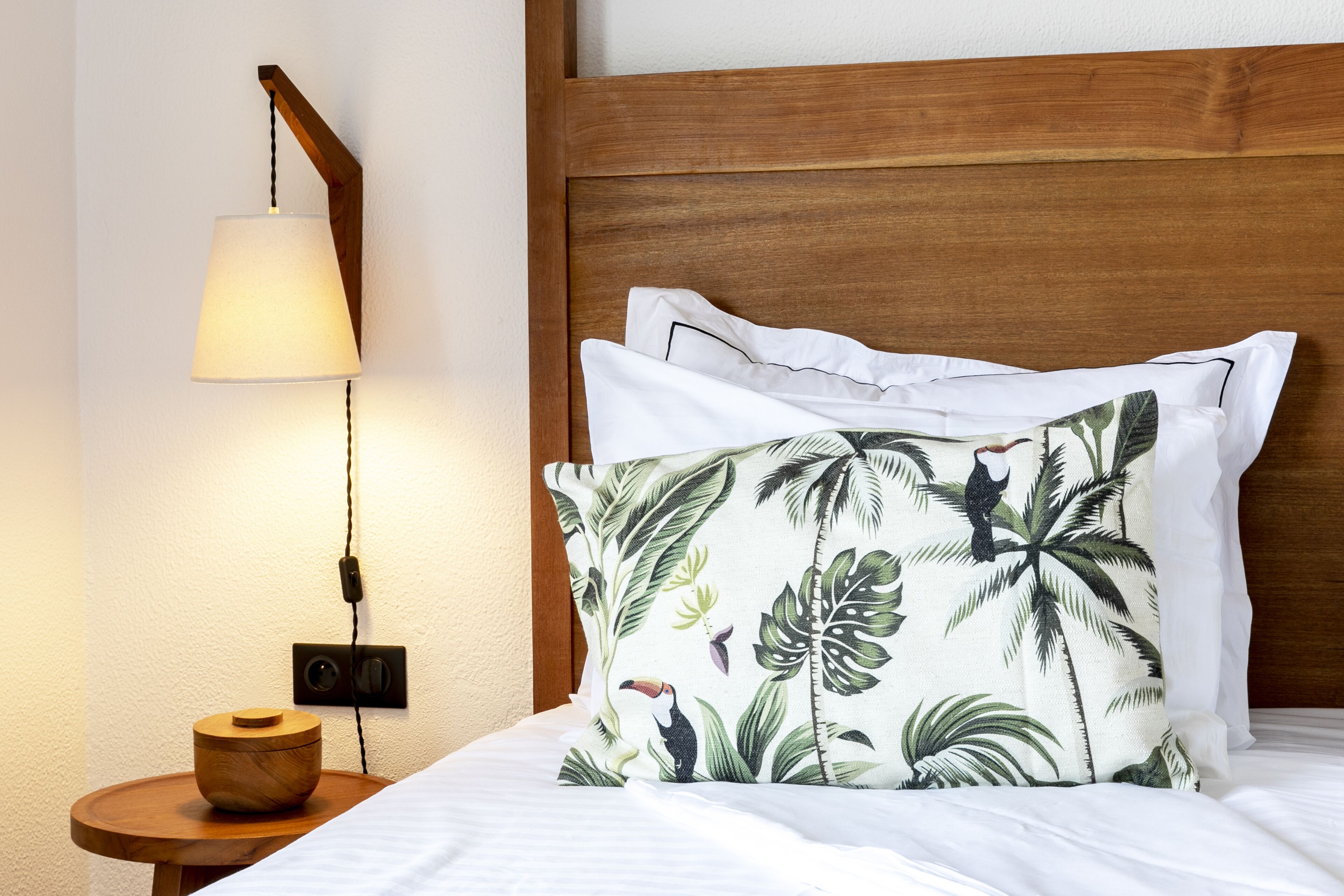 Un lit bien fait, avec une literie blanche, une tête de lit en bois et un coussin aux motifs tropicaux.