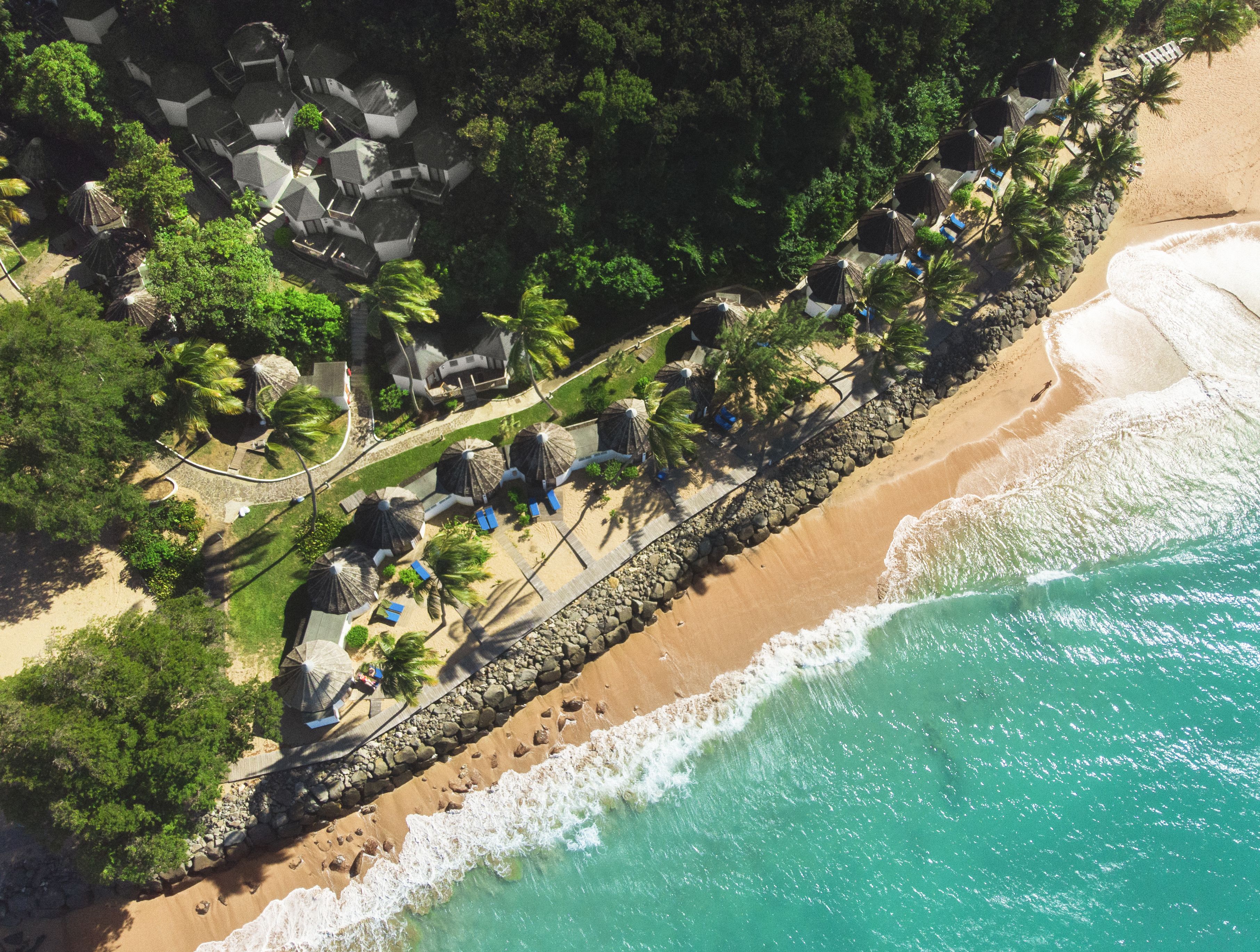 An aerial view of a beach resort with multiple bungalow-style accommodations, a rocky shoreline, and a clear turquoise sea.