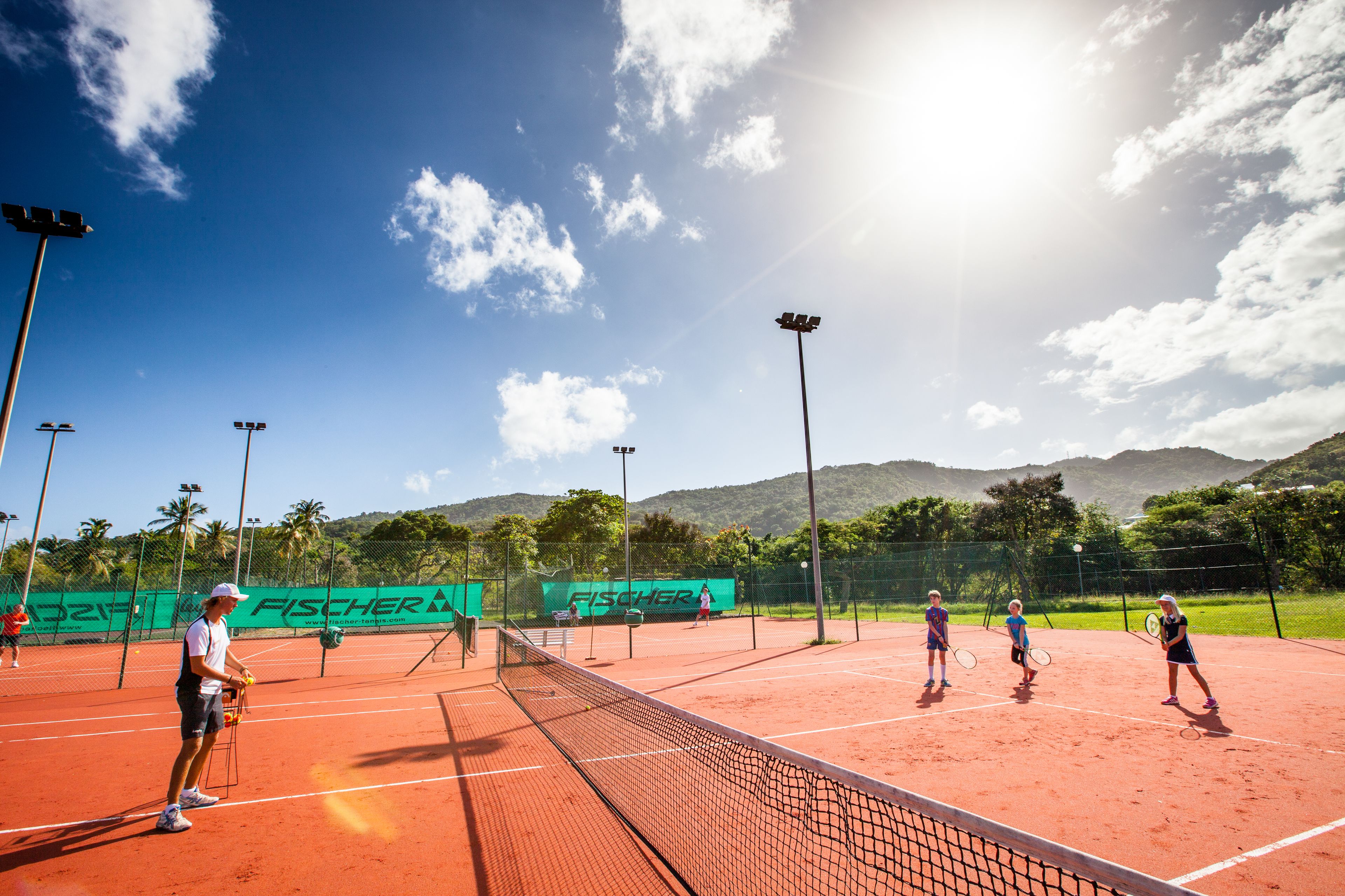 A tennis court with a net, surrounded by a fence, and a clear sky with clouds.