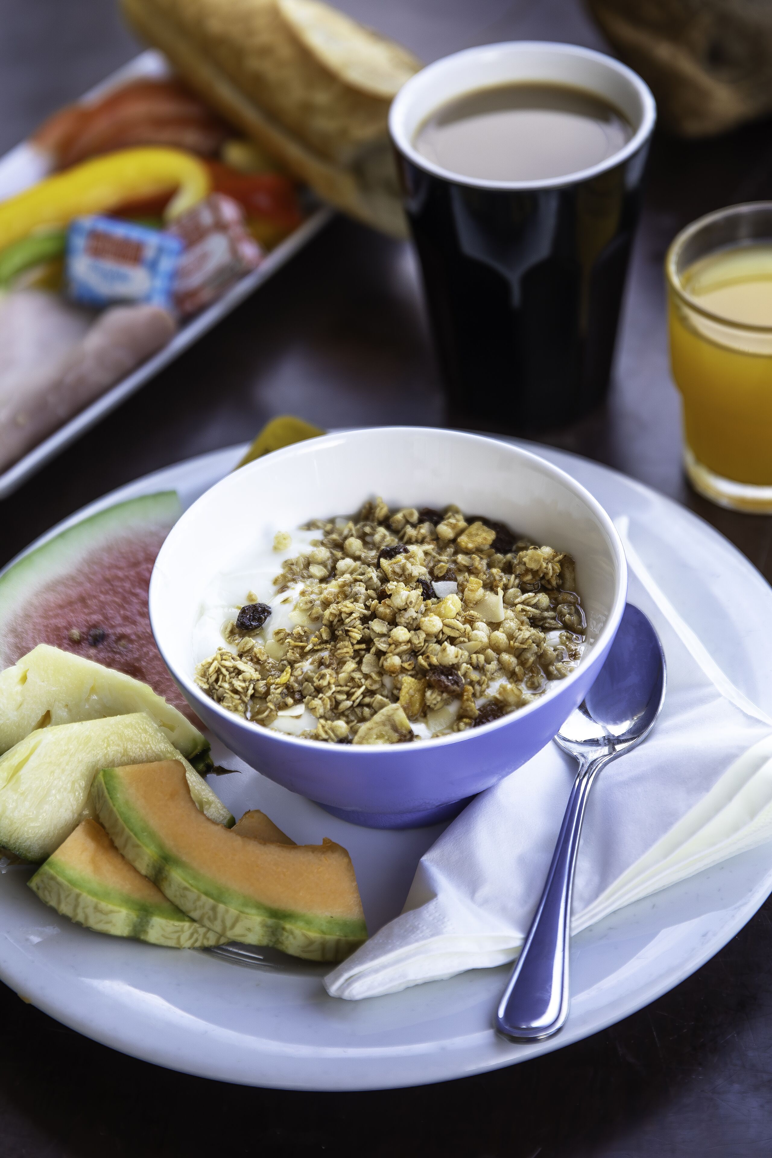 Un plato de desayuno con yogur, granola, rodajas de melón y una taza de café.