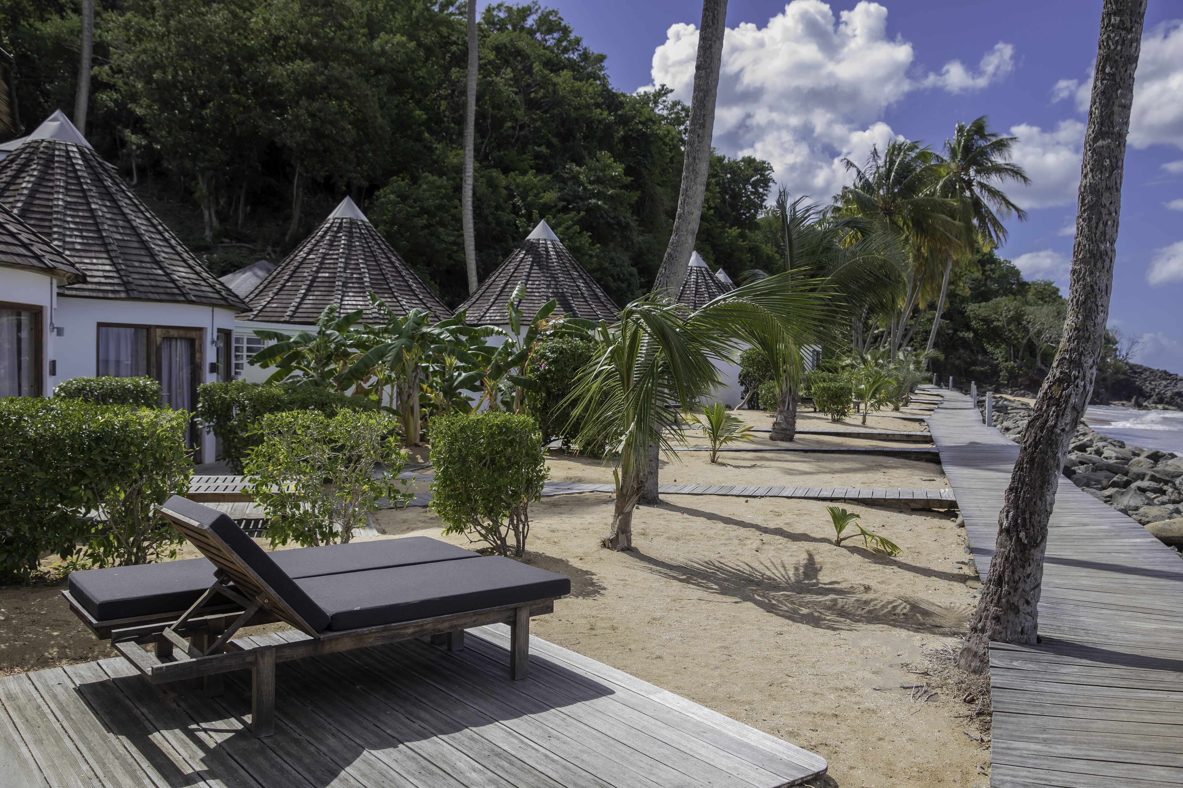 A wooden deck with lounge chairs, a sandy beach, palm trees, and a row of buildings with peaked roofs.
