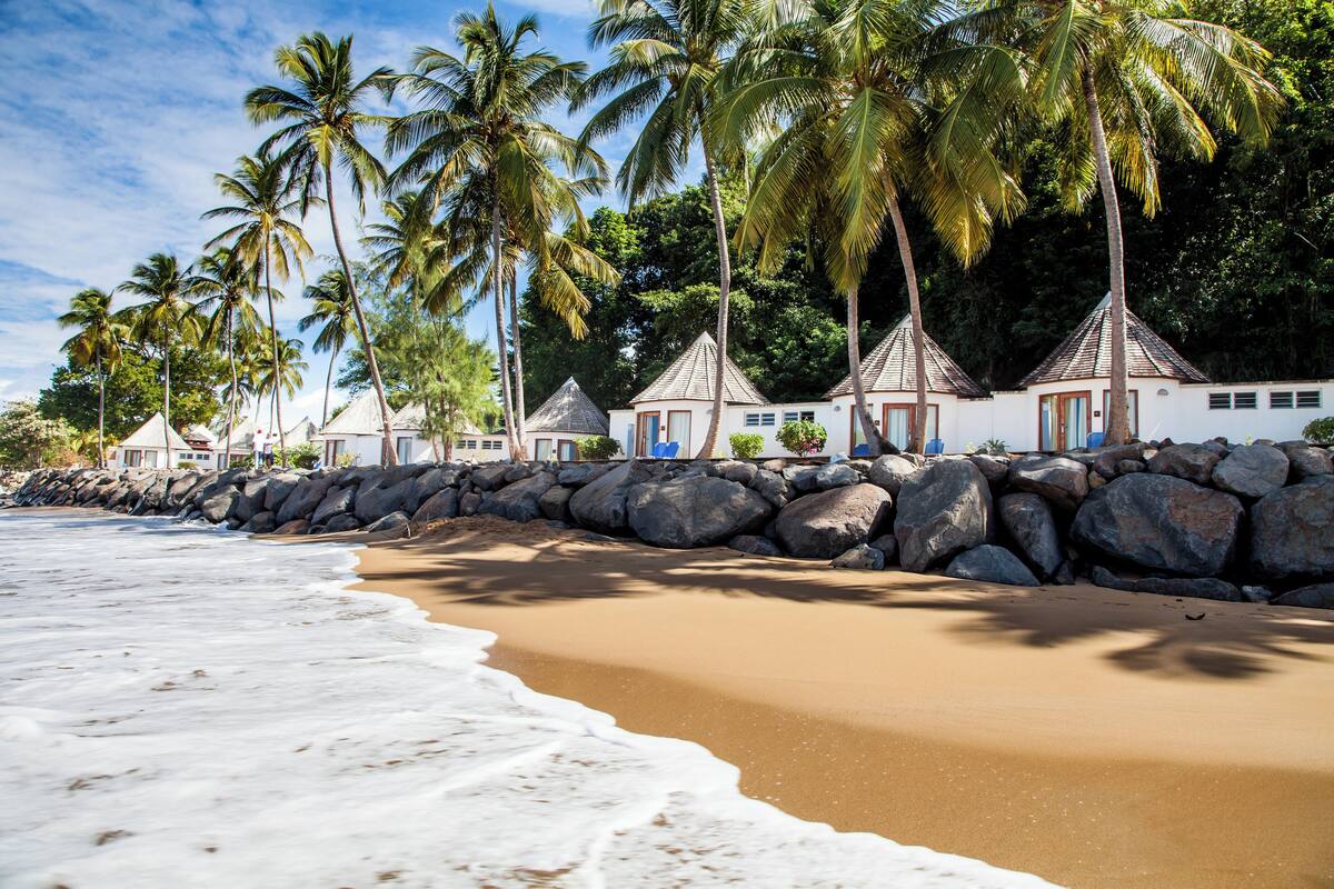 Ein Strand mit einer Reihe weißer Gebäude mit roten Dächern, Palmen und einer felsigen Küstenlinie.