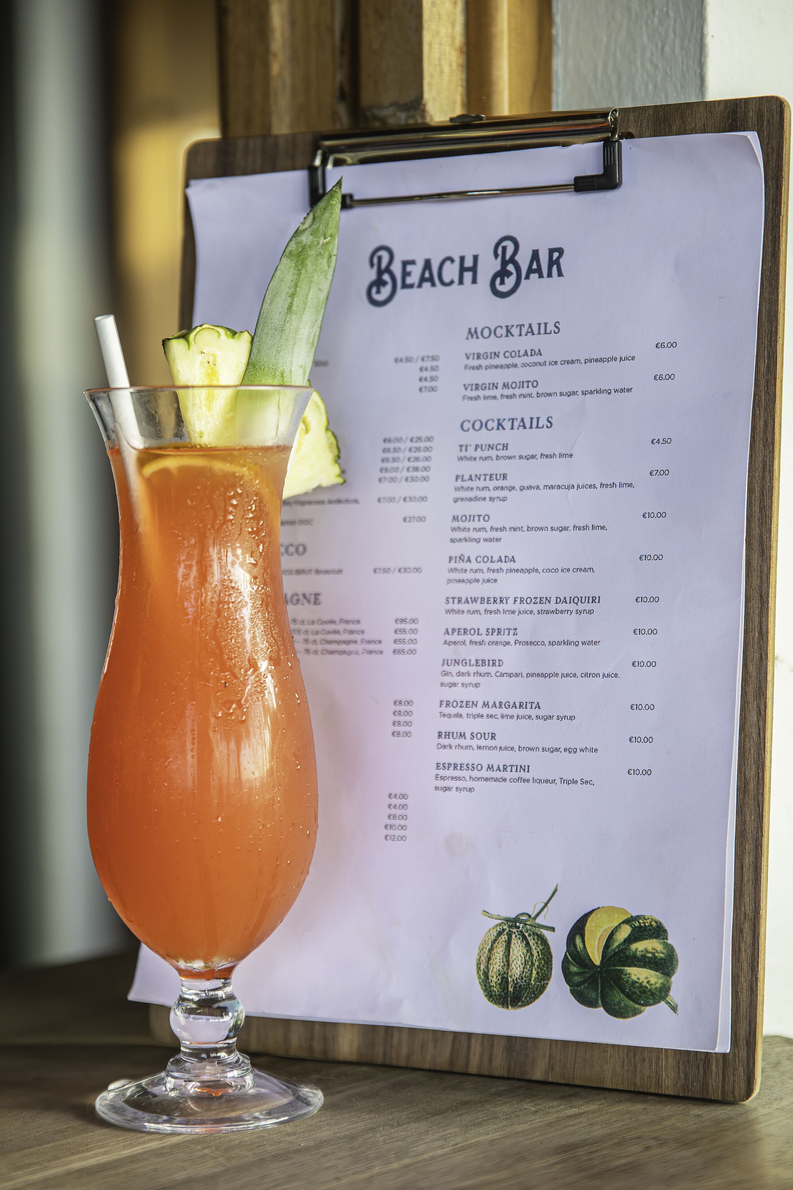 A cocktail in a glass with a menu for 'Beach Bar' behind it listing mocktails and cocktails with prices.