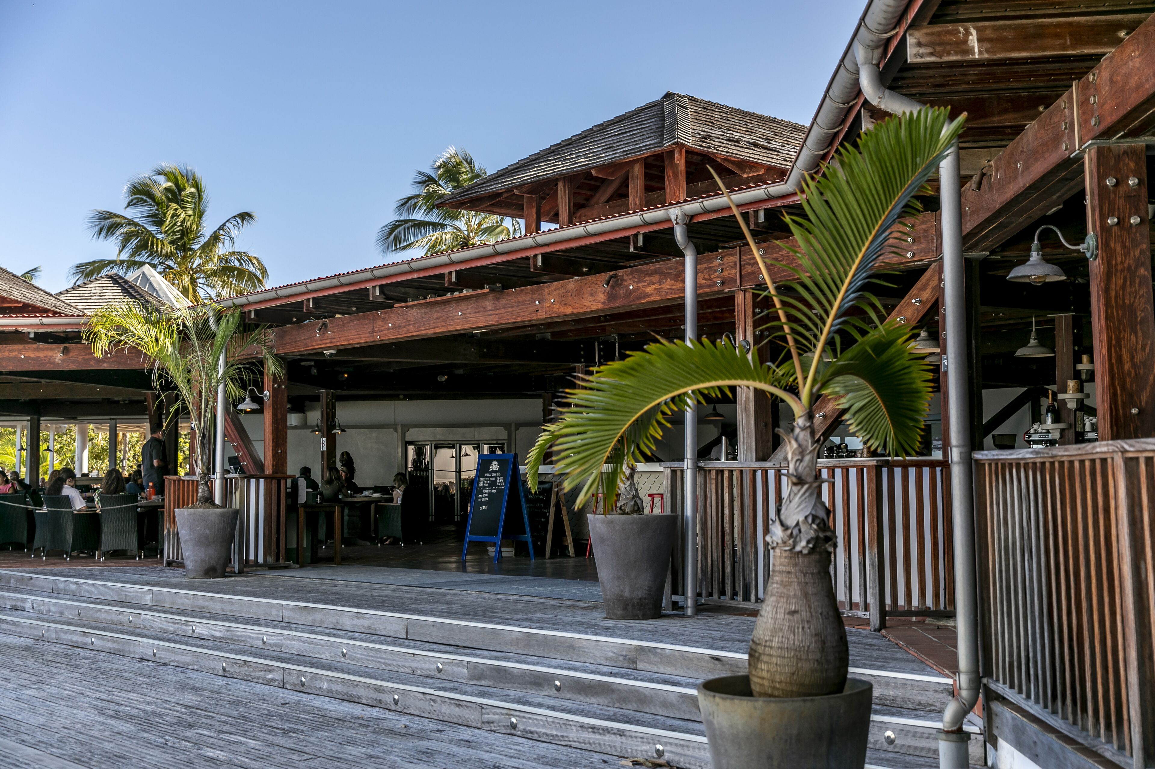An outdoor dining area with wooden deck, large potted plants, and a covered seating area.