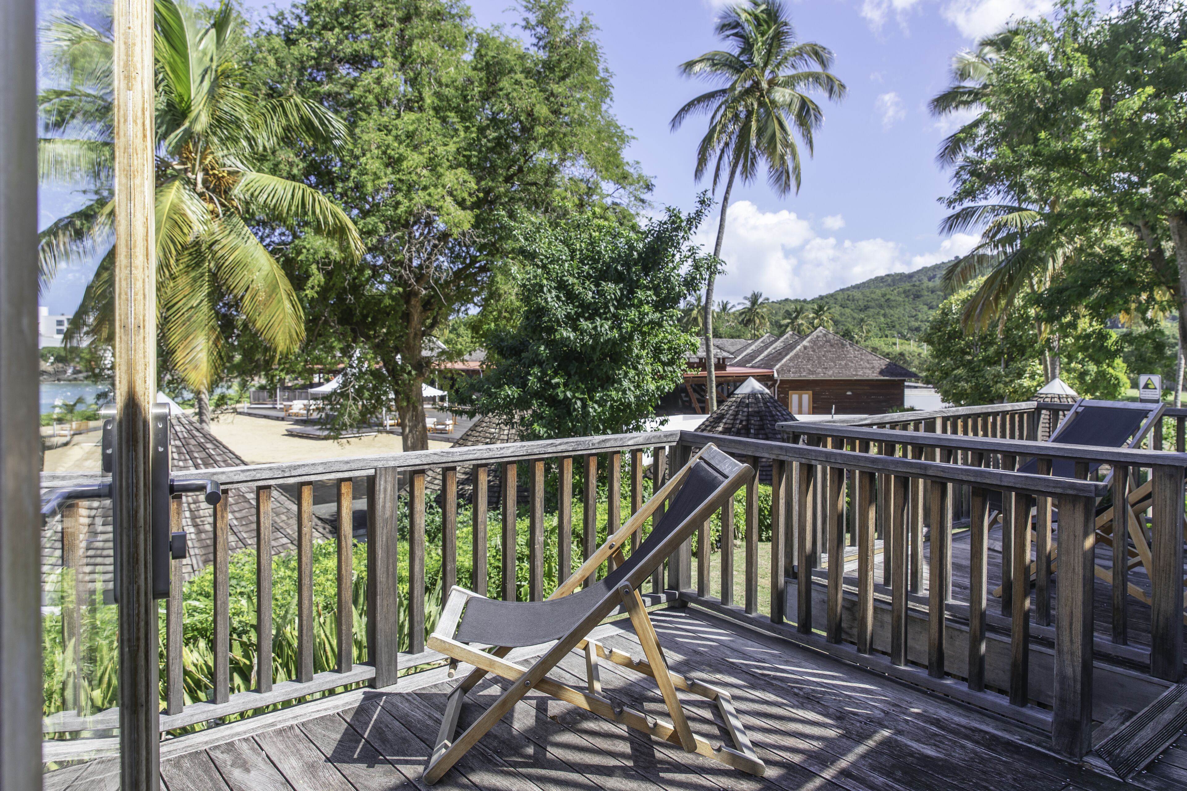 A wooden deck with a single lounge chair, surrounded by palm trees and a view of a beach and buildings in the background.