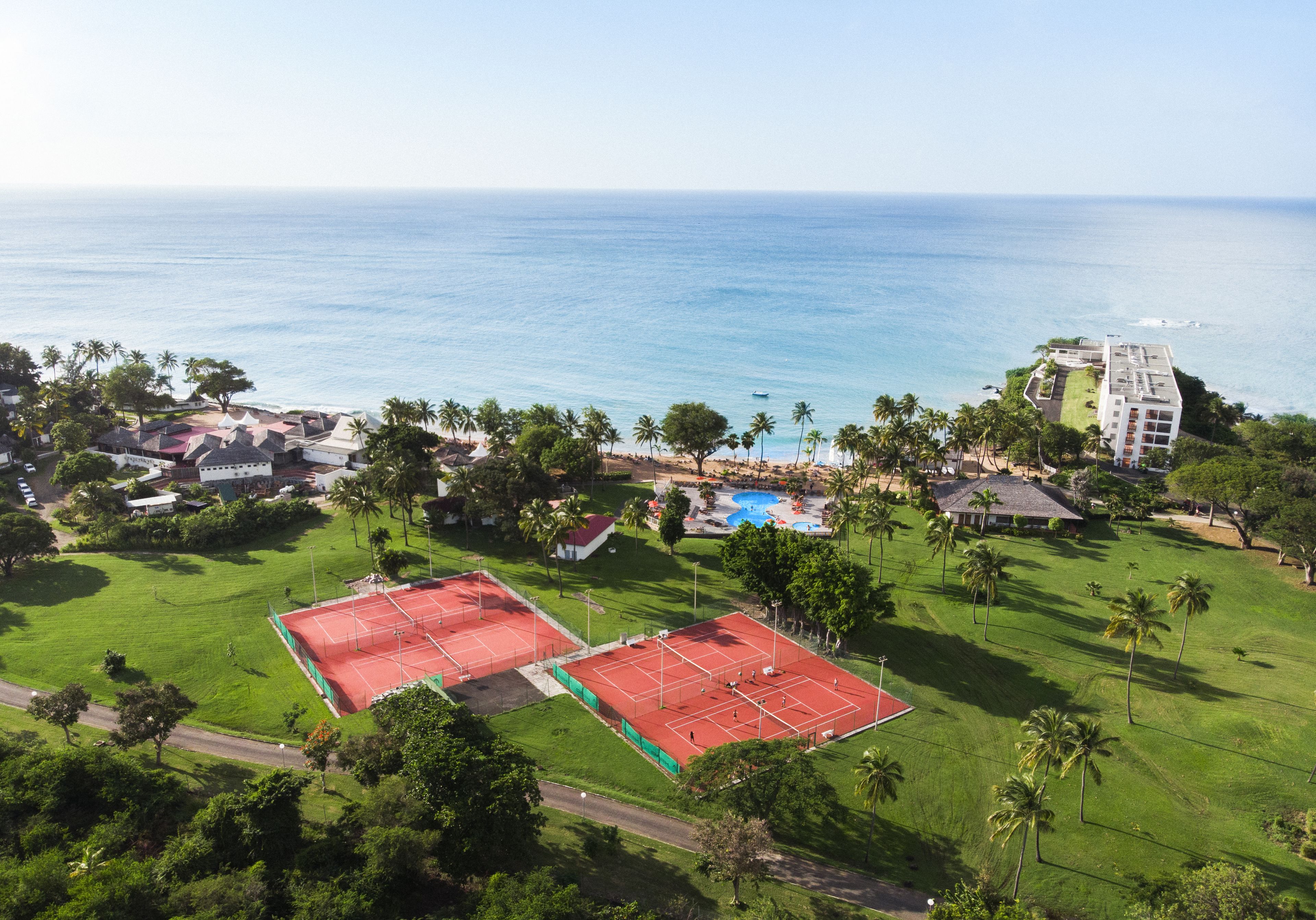 An aerial view of a coastal area with two tennis courts, a swimming pool, and a hotel.