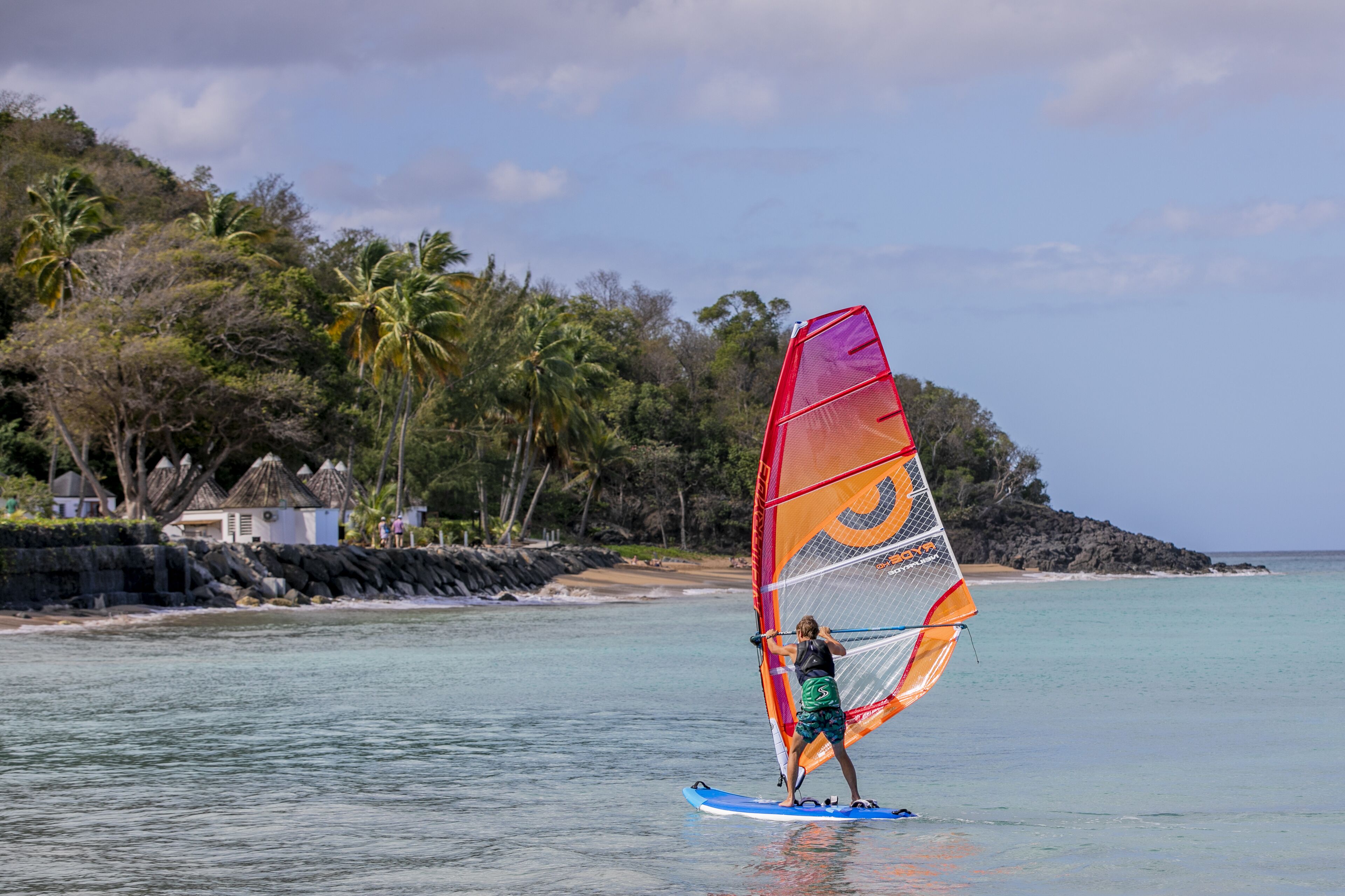 A person windsurfing on a beach with palm trees and a rocky shoreline in the background.