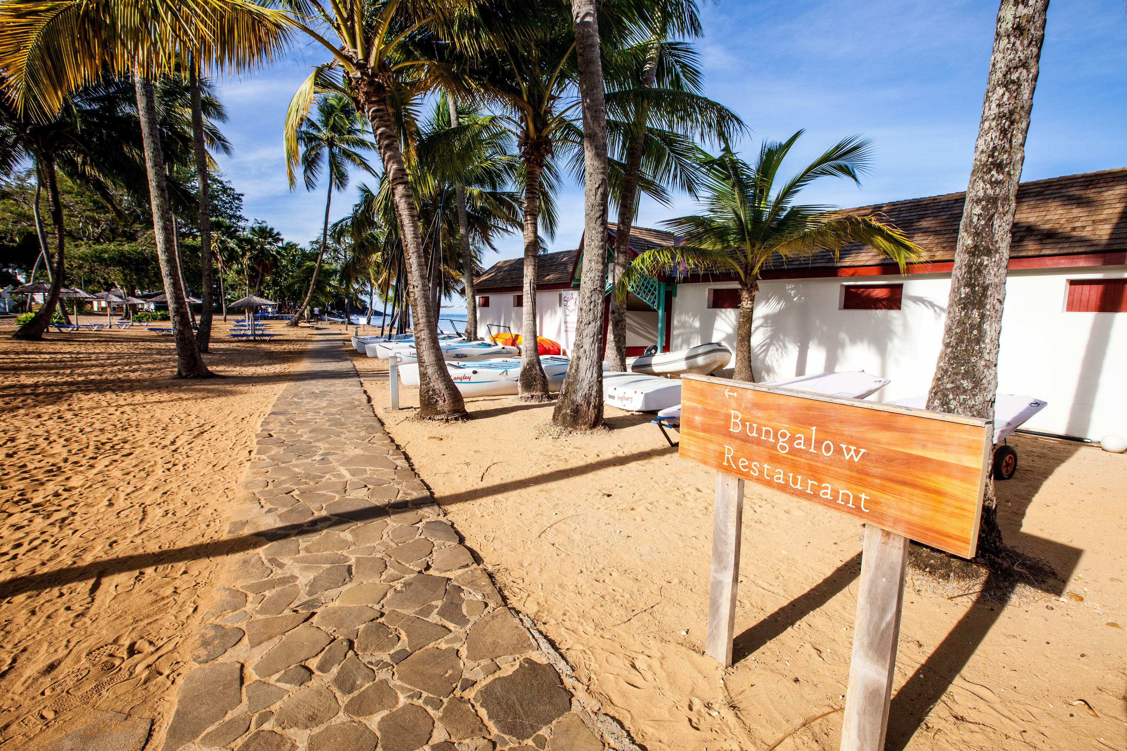 Un restaurante frente al mar con un letrero, palmeras y sombrillas de playa.
