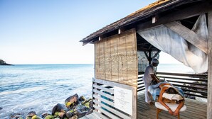 A wooden structure with a bamboo wall and a wooden deck extending over the water, featuring a massage chair and a person receiving a massage. - Langley Resort Fort Royal (Deshaies)