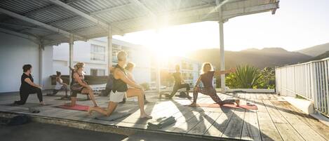 A group of people performing yoga on a wooden deck under a metal roof with a view of mountains in the background.