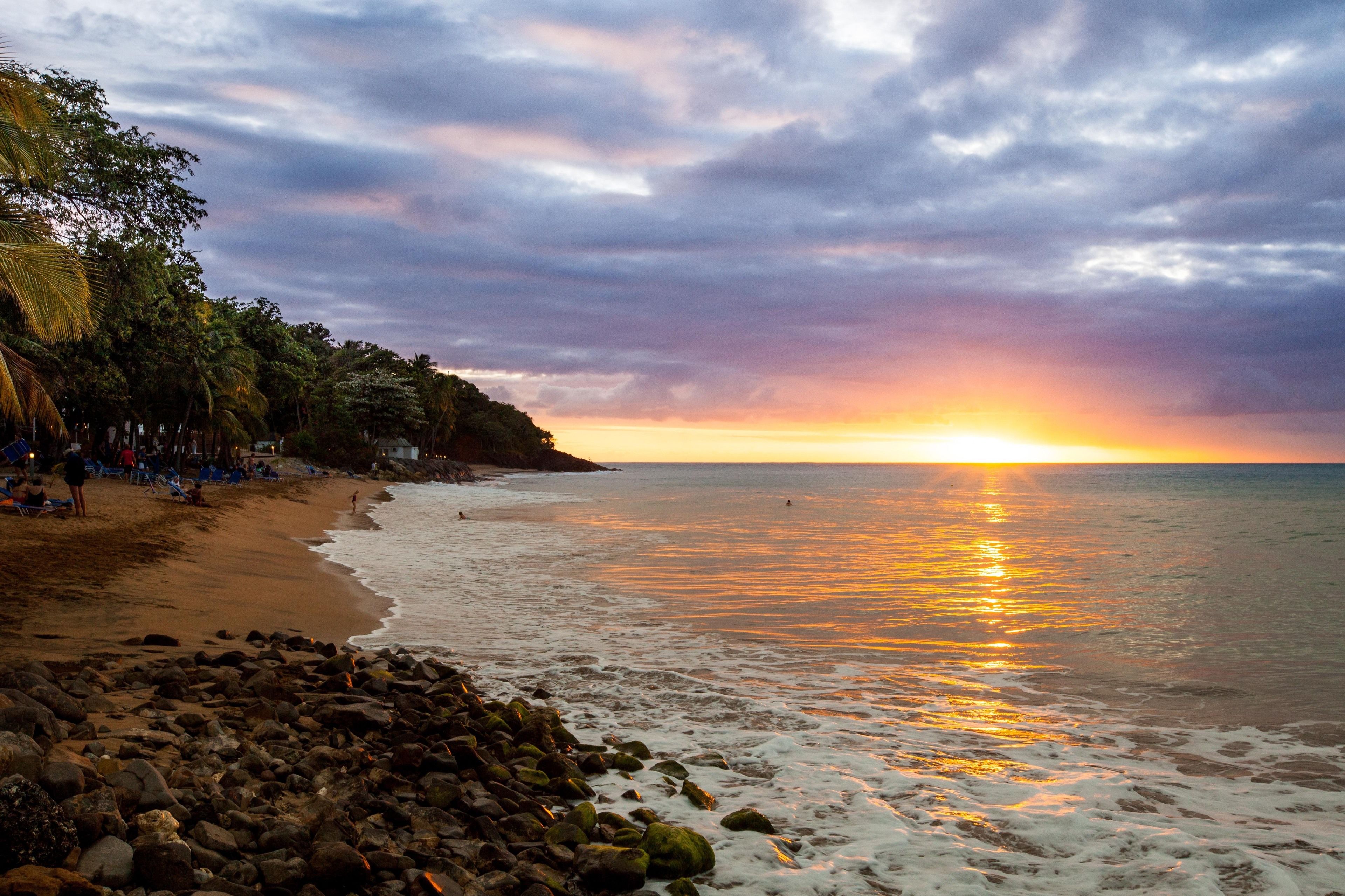 Ein Strand mit Sonnenuntergang, felsiger Küste und Palmen.