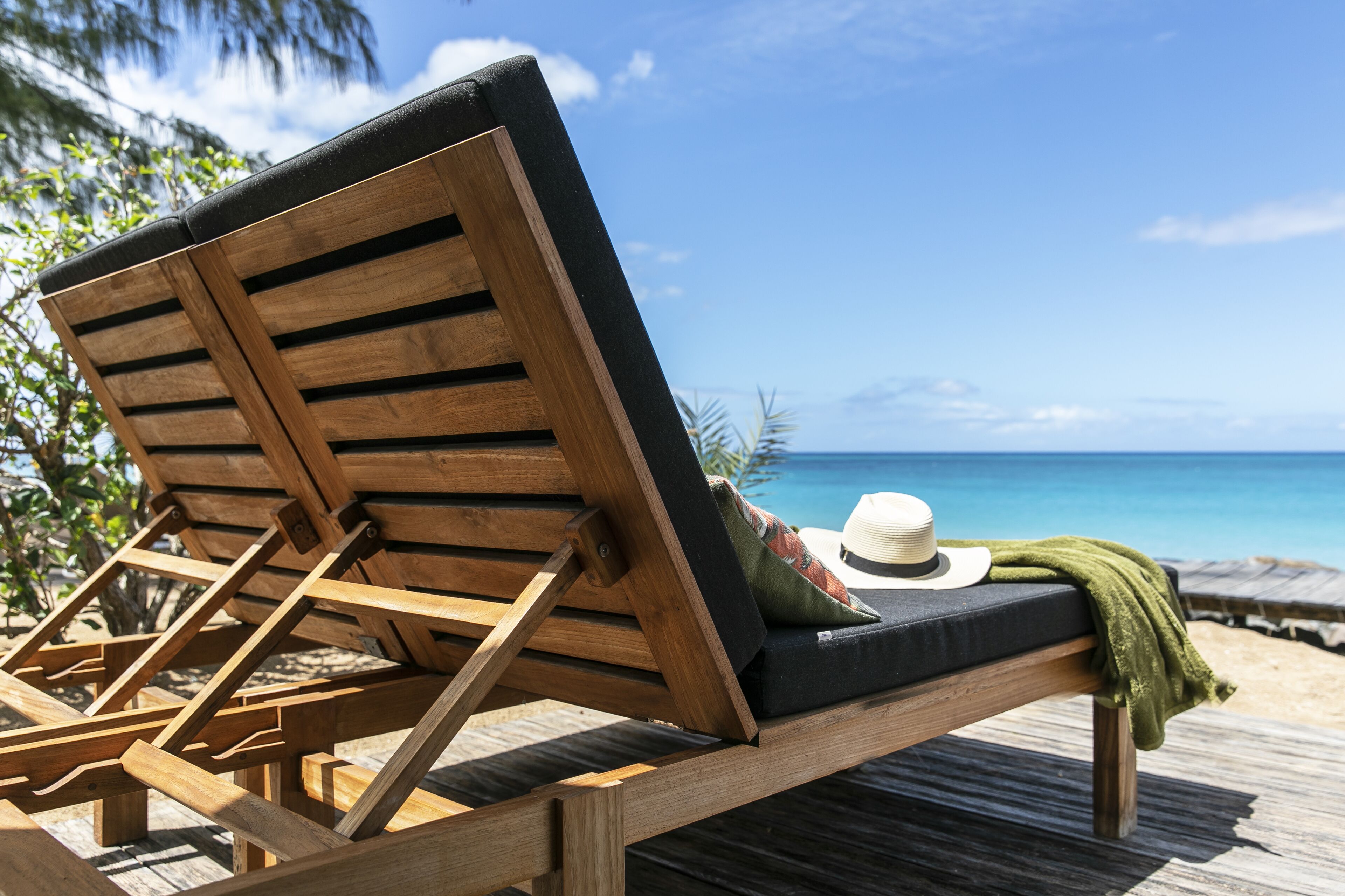 A wooden lounge chair with a cushion and a hat on it, positioned on a wooden deck with a view of the ocean.