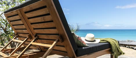 A wooden lounge chair with a cushion and a hat on it, positioned on a wooden deck with a view of the ocean.