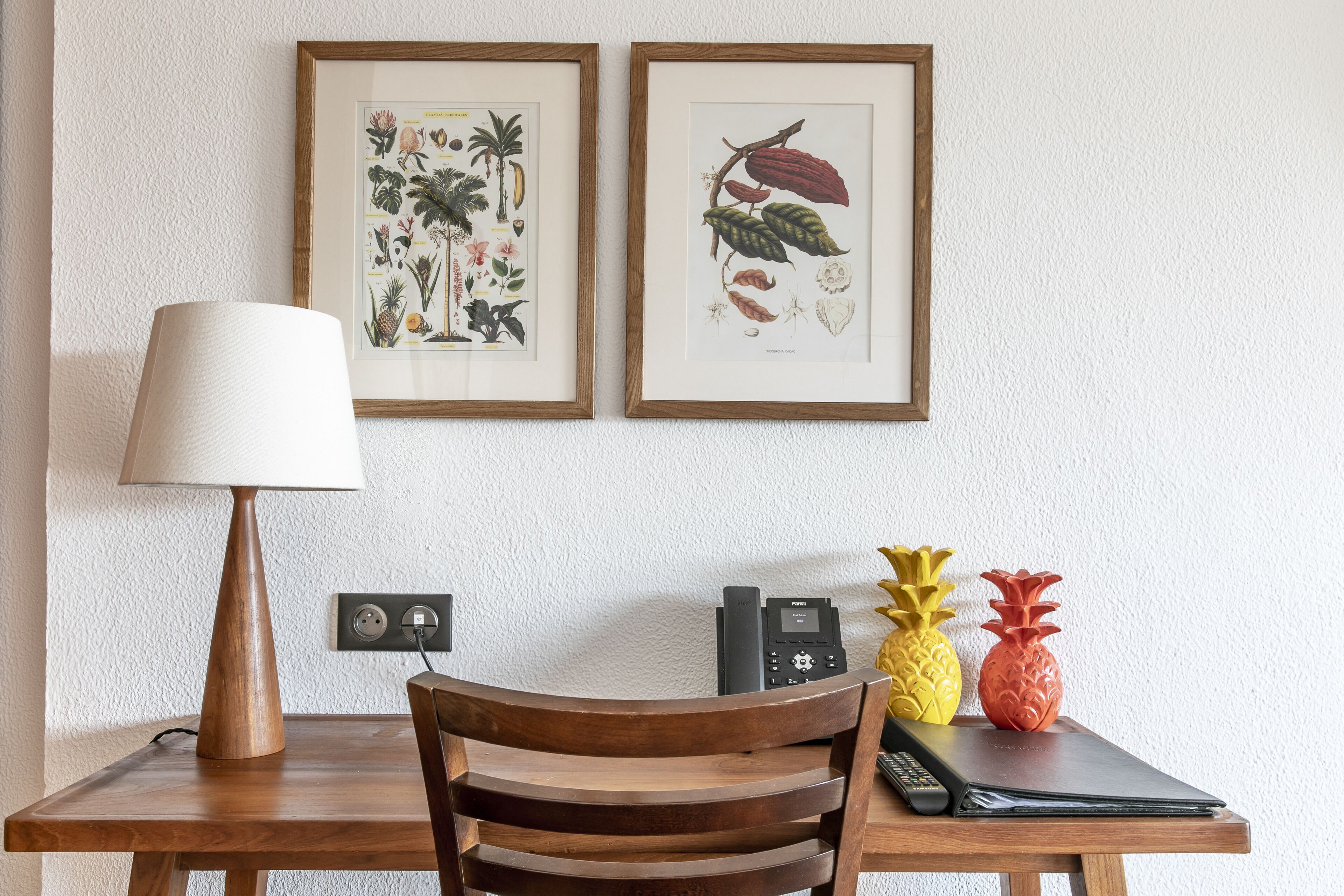 A wooden table with a lamp, framed botanical prints, and two pineapple-shaped vases.