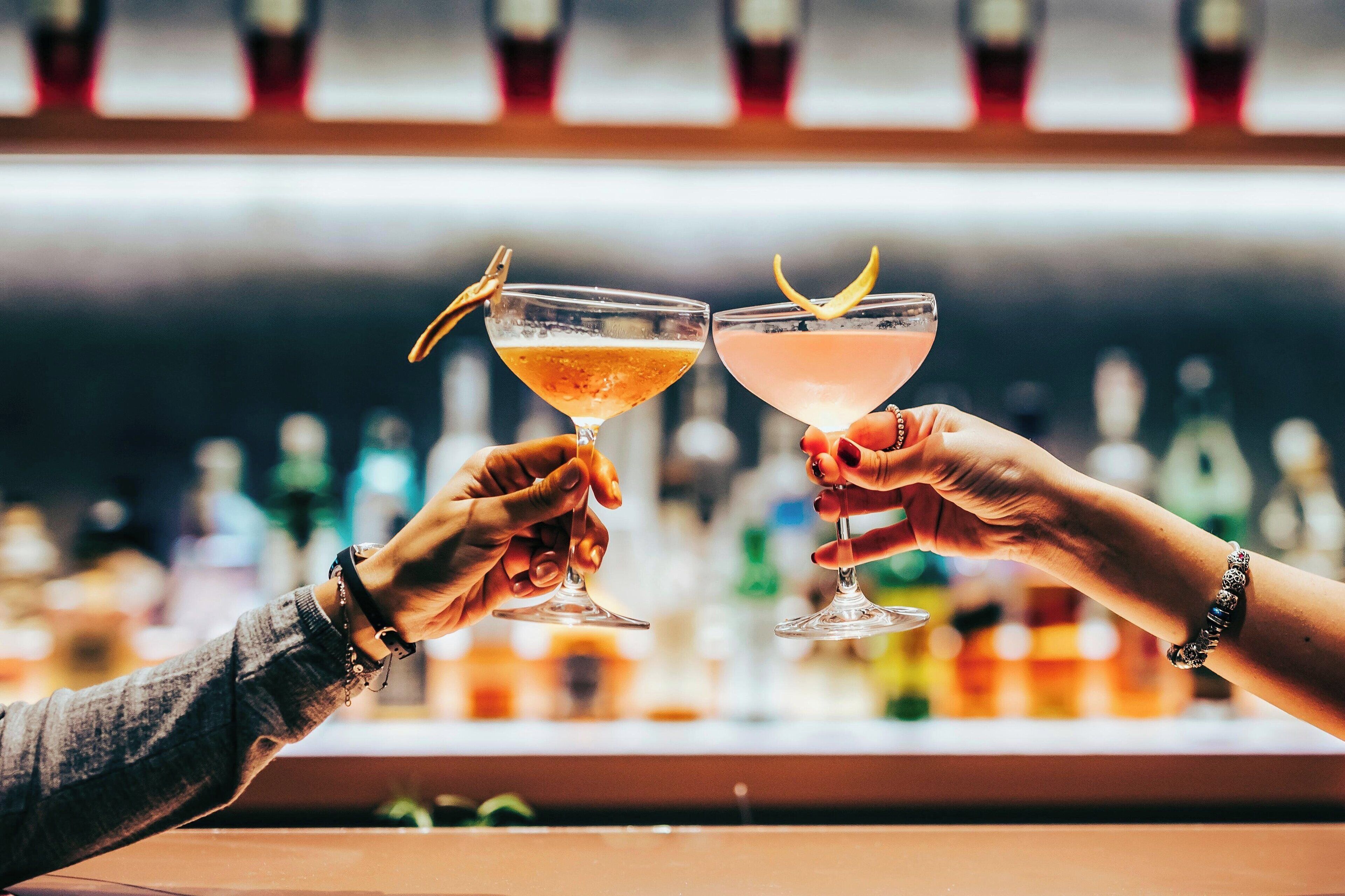 Duas mãos segurando copos de coquetel com guarnições, em um bar com garrafas ao fundo.