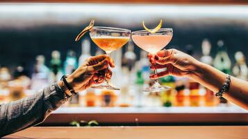 Duas mãos segurando copos de coquetel com guarnições, em um bar com garrafas ao fundo.