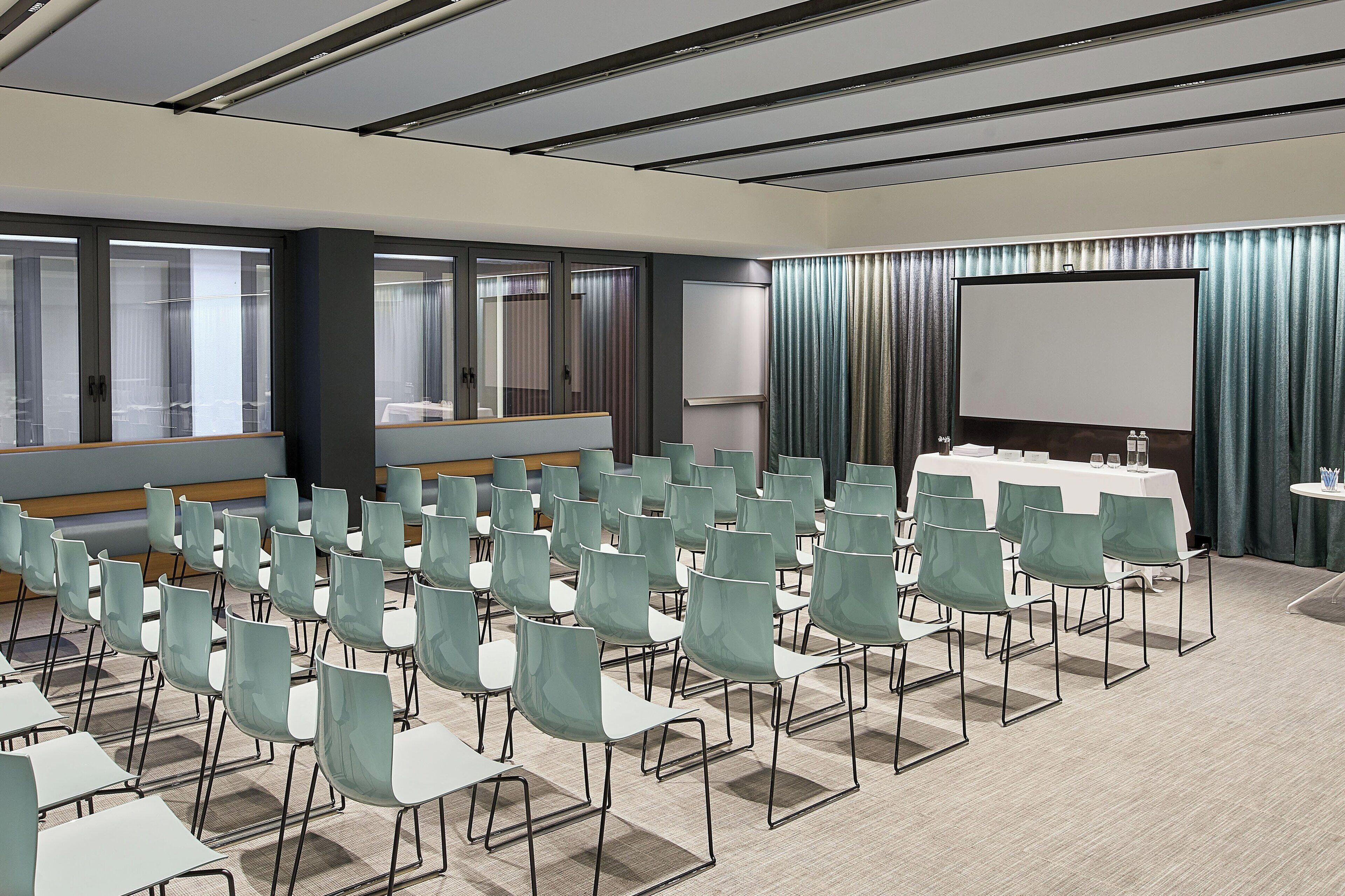 Una sala conferenze con file di sedie e tavoli, ampie finestre con tende e uno schermo di proiezione.