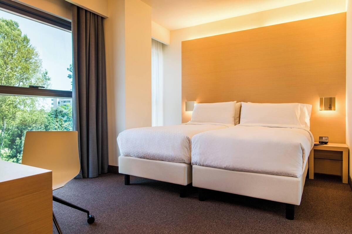 Een tweepersoonsbed met houten hoofdbord, een nachtkastje met lamp, en uitzicht op bomen door een raam.