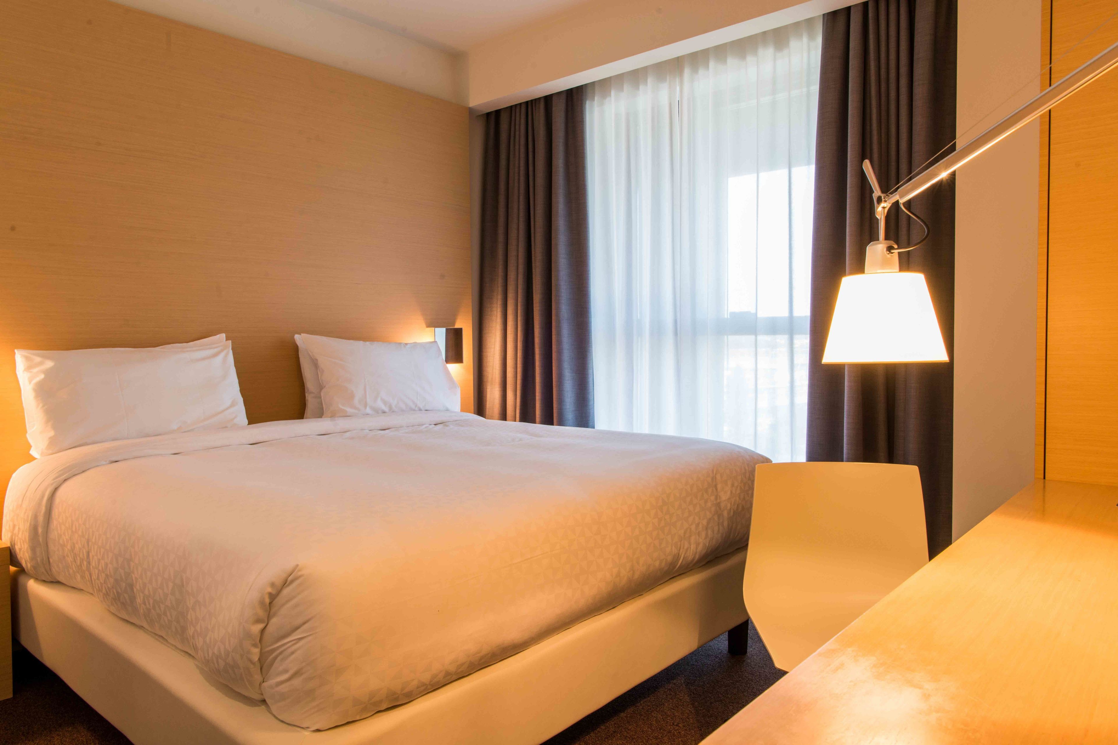 Hotellihuone, jossa on suuri sänky, yöpöytä, lamppu ja ikkunasta avautuva näkymä kaupunkiin.