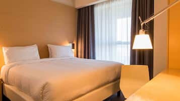 A hotel room with a large bed, a bedside table, a lamp, and a view of the city through the window.