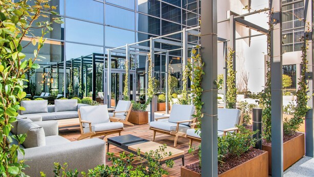 Un espacio al aire libre moderno con sofás y sillones, rodeado de follaje y un frente de cristal.