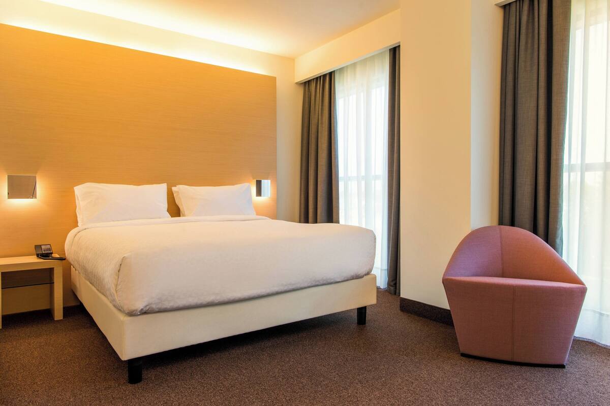 Een hotelkamer met een bed, nachtkastje, stoel en een groot raam met gordijnen.