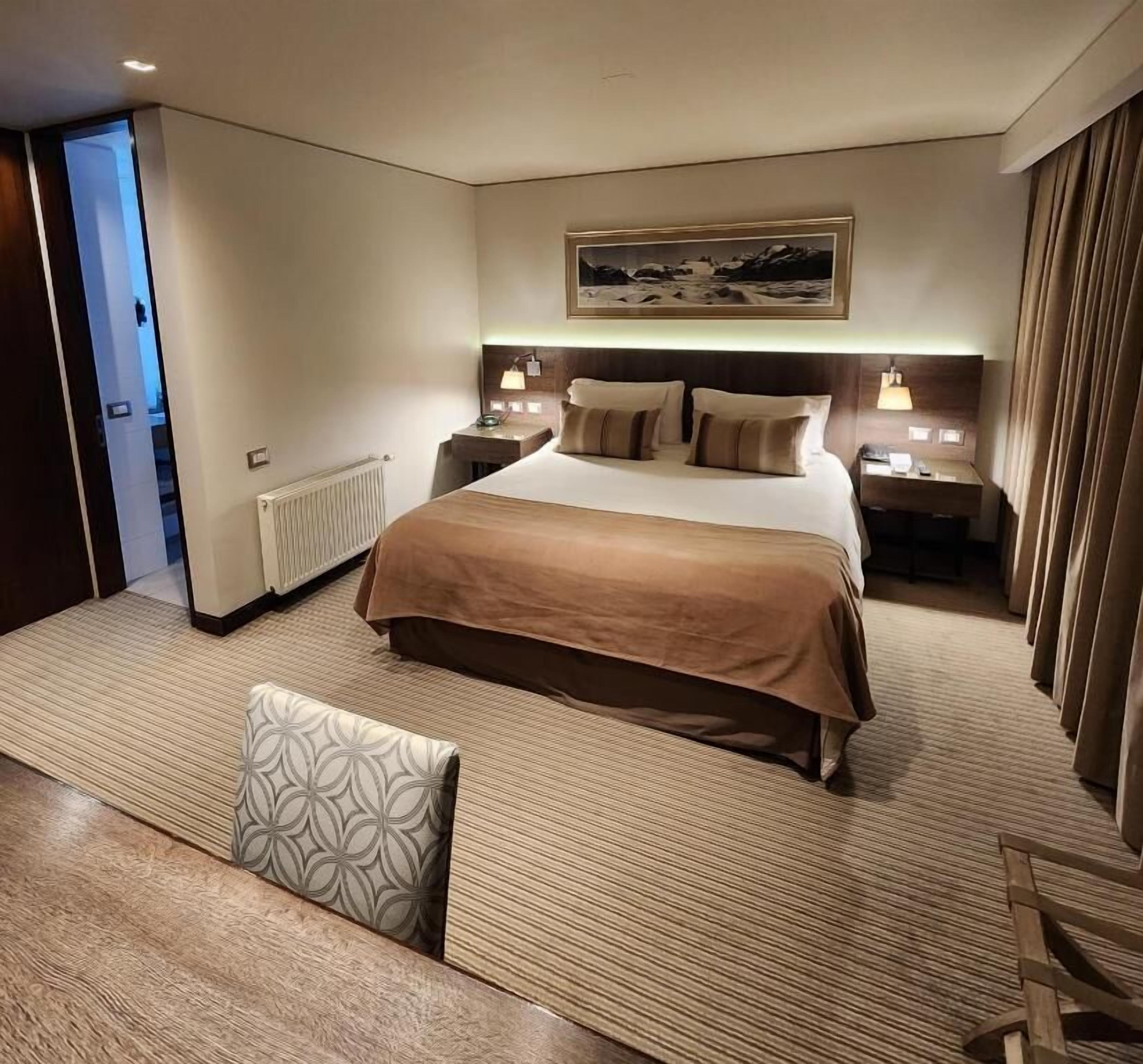 A hotel room with a large bed, bedside tables, a nightstand, a window with curtains, and a view of a snowy landscape.
