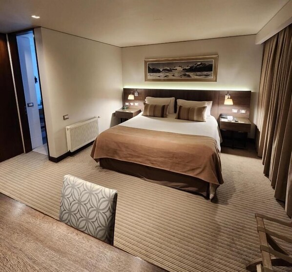 A hotel room with a large bed, bedside tables, a nightstand, a window with curtains, and a view of a snowy landscape.