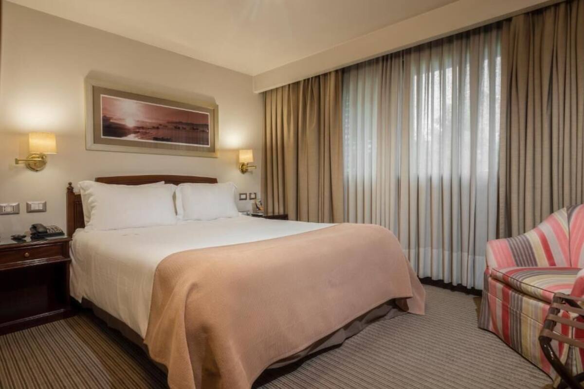 A hotel room with a bed, a chair, a painting on the wall, and curtains.