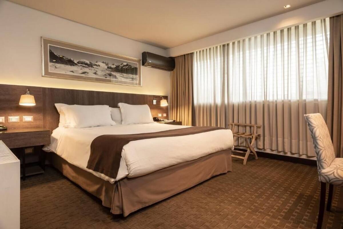 A hotel room with a large bed, two bedside tables, a chair, a picture on the wall, and a window with curtains.