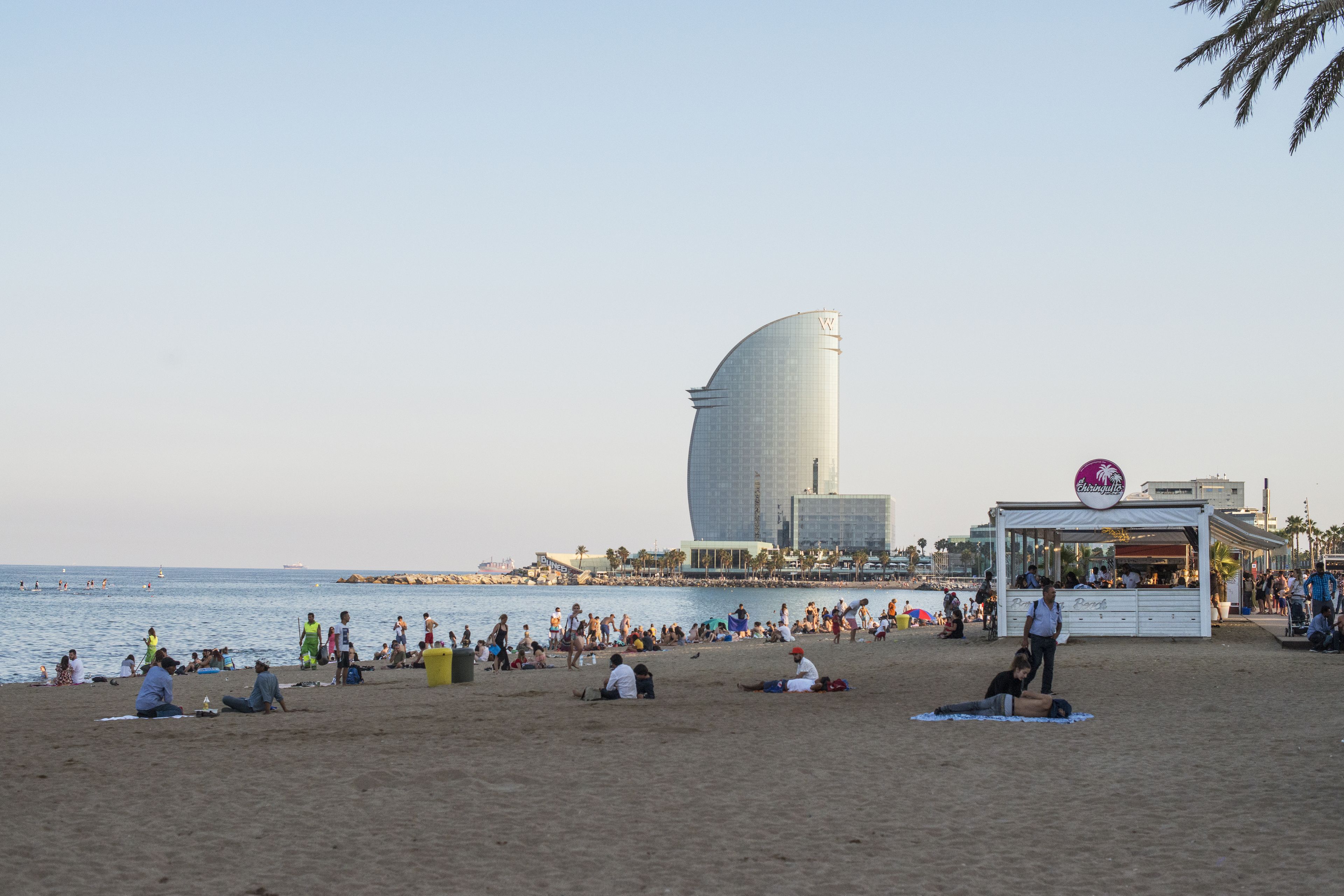 A beach with people relaxing, a large building, and a food kiosk.