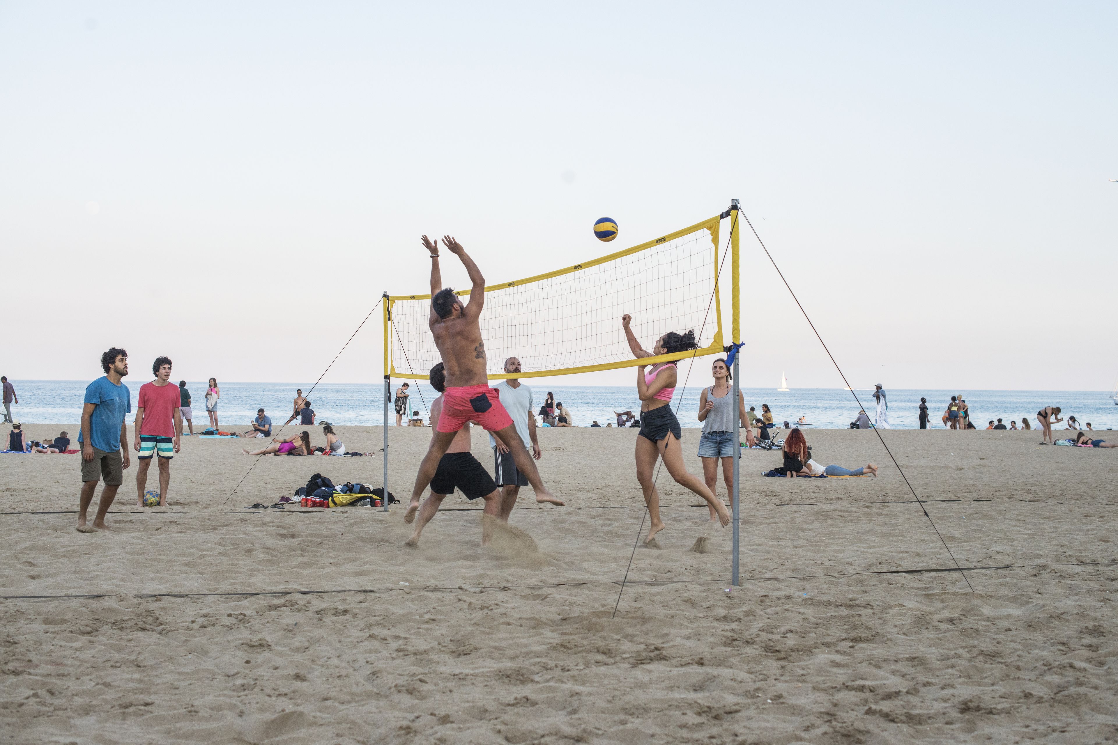 A beach volleyball game with players in action, spectators on the sand, and a clear view of the ocean in the background.