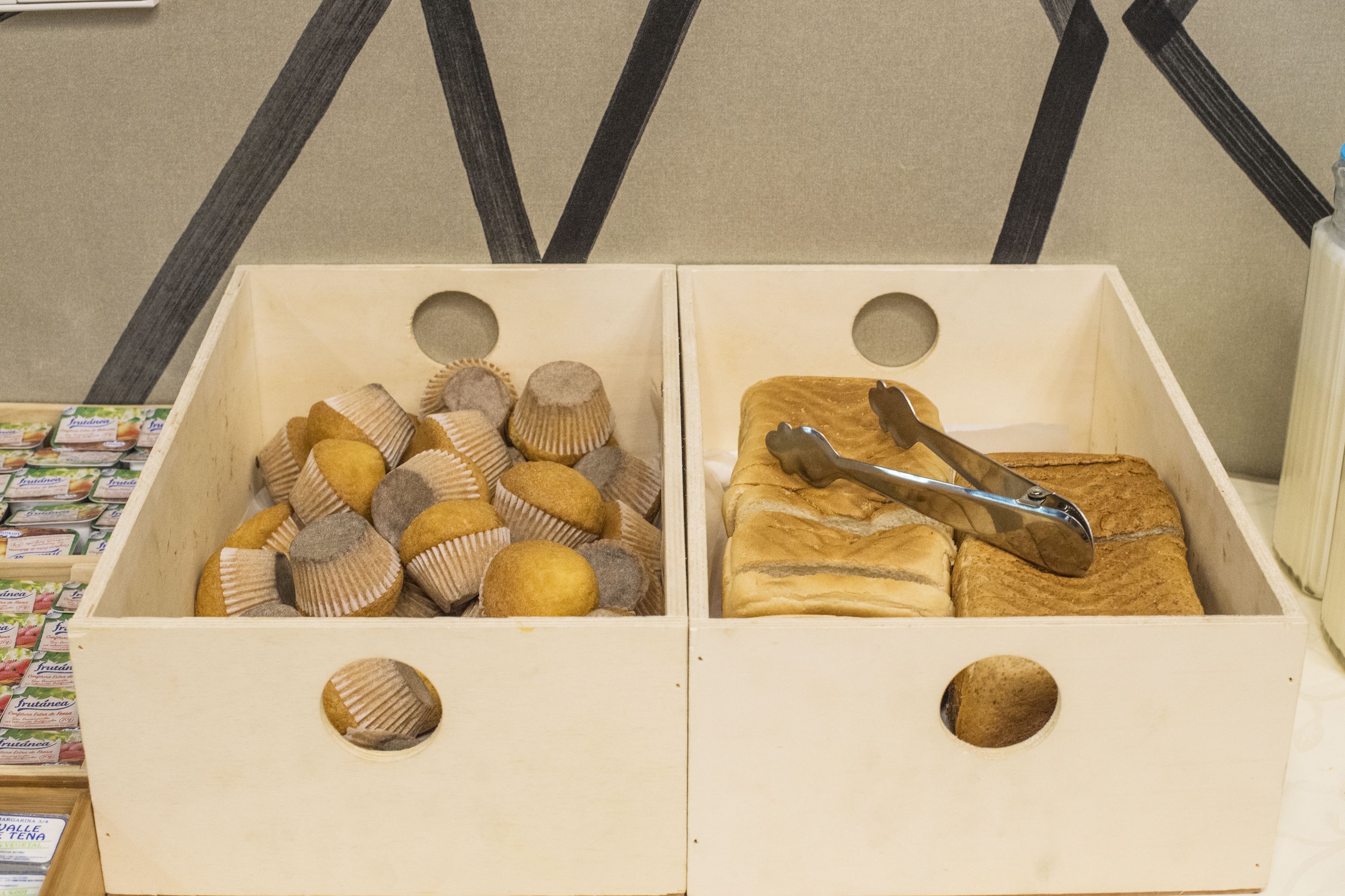 Two wooden boxes with circular cutouts, one filled with round, brown objects and the other with a pair of tongs resting on a loaf of bread.