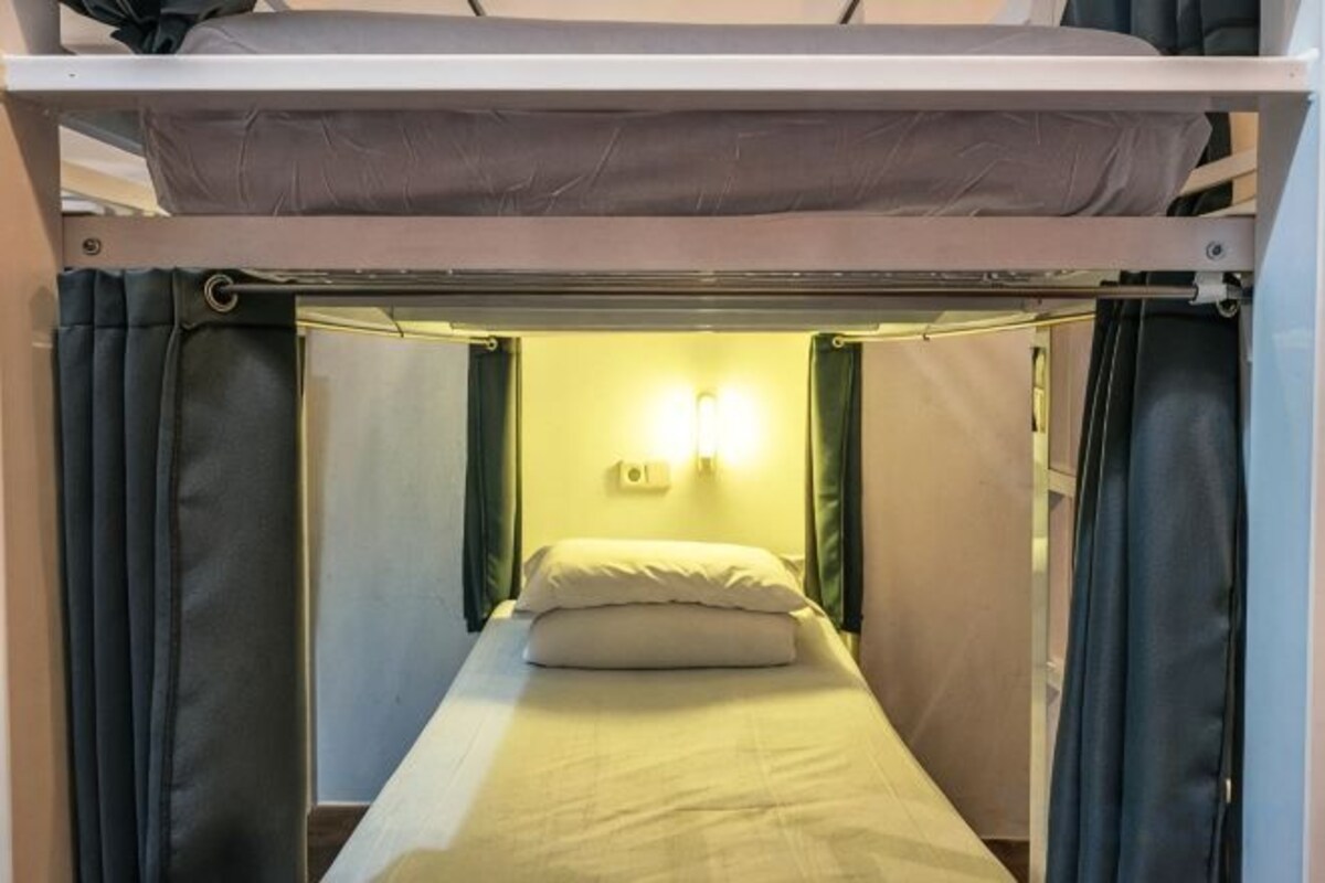 A bunk bed with a single bed below, curtains on either side, and a wall-mounted light fixture.