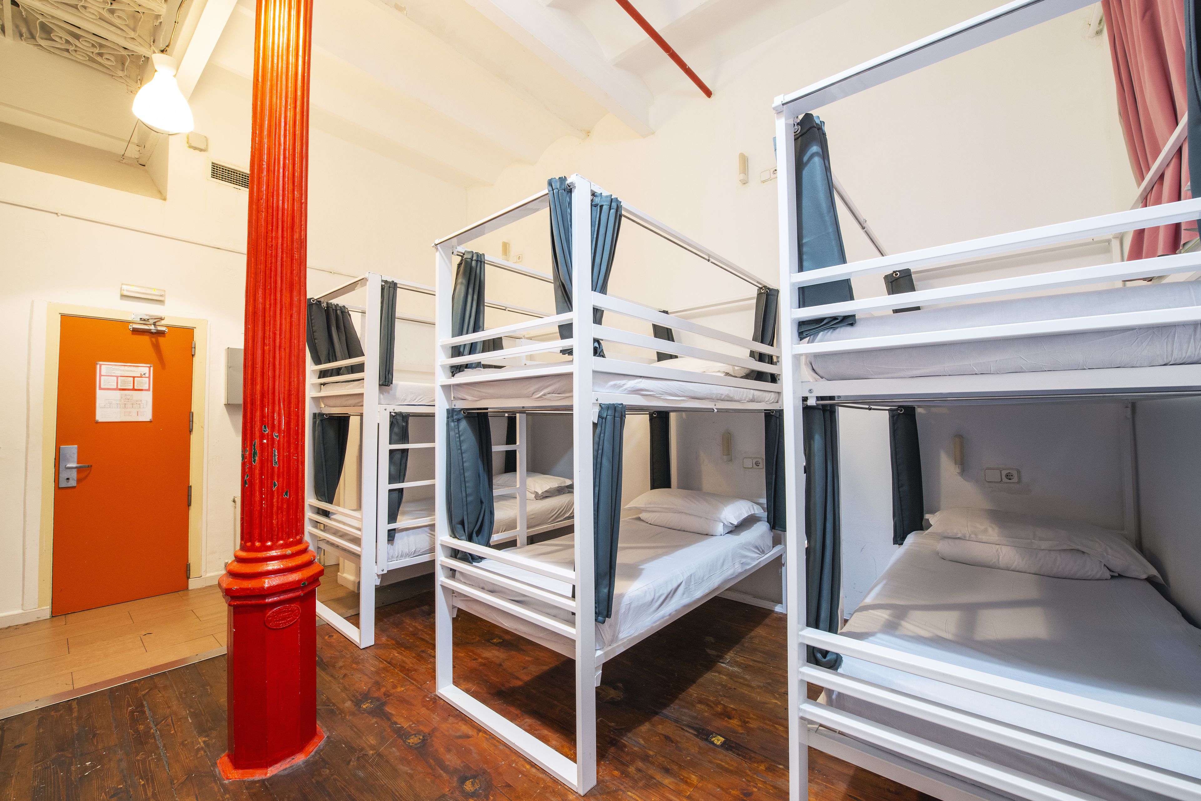 A dormitory room with bunk beds, a red pillar, and a door with an emergency exit sign.