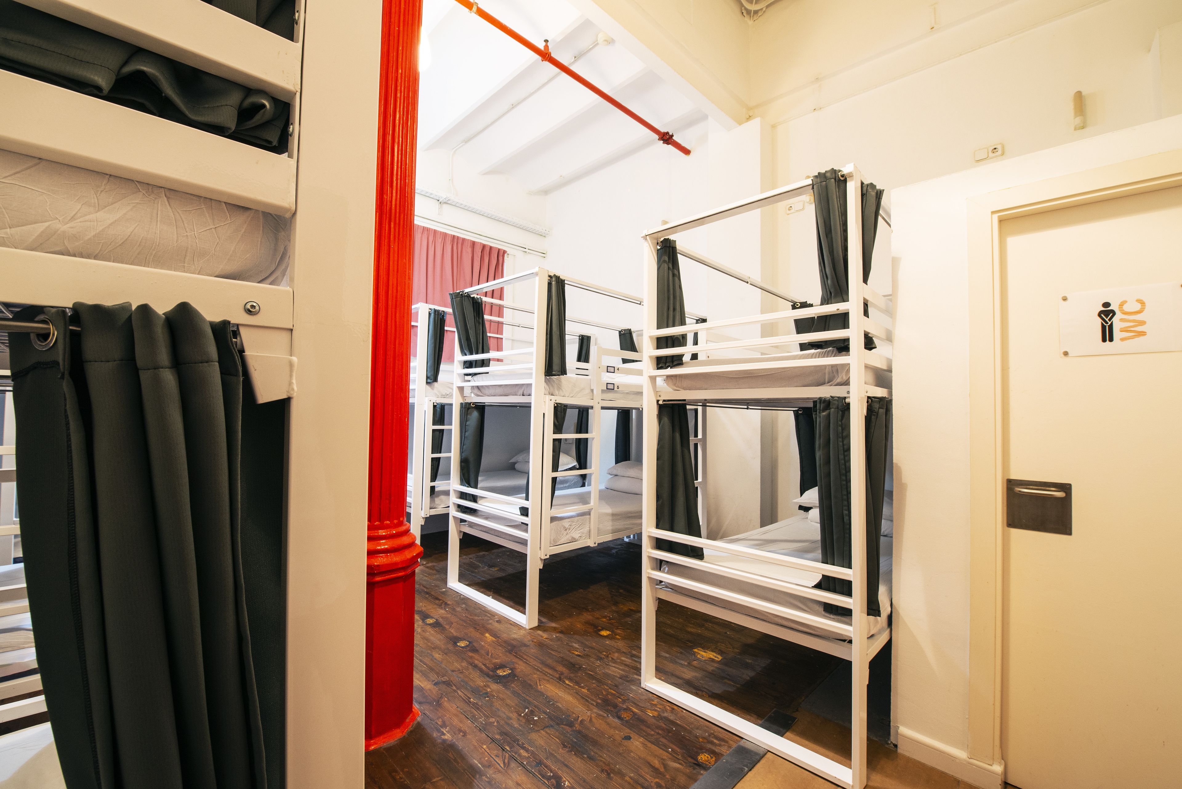 A dormitory room with bunk beds, a red curtain, and a door with a WC sign.