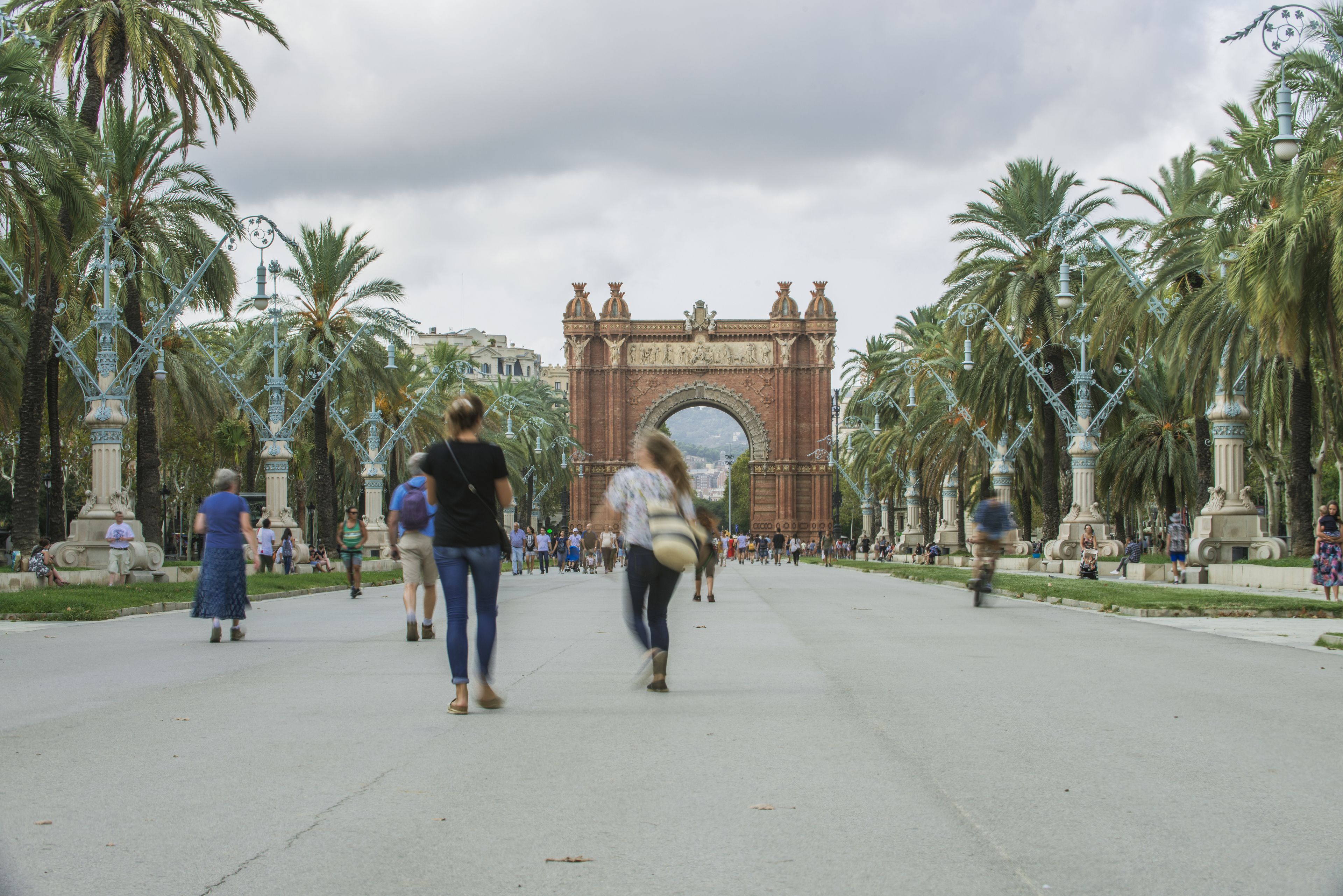 A wide pedestrian pathway lined with palm trees and decorative street lamps, leading to a large, ornate archway.