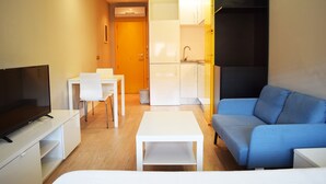 Un salon moderne avec un téléviseur à écran plat, une table basse blanche, un canapé bleu, une table à manger avec des chaises, et un coin cuisine équipé d’un réfrigérateur et de placards.