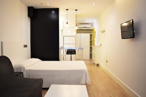 Ein Hotelzimmer mit zwei Betten, einem kleinen Tisch, einem Stuhl, einem an der Wand befestigten Fernseher und einer Küchenzeile im Hintergrund.
