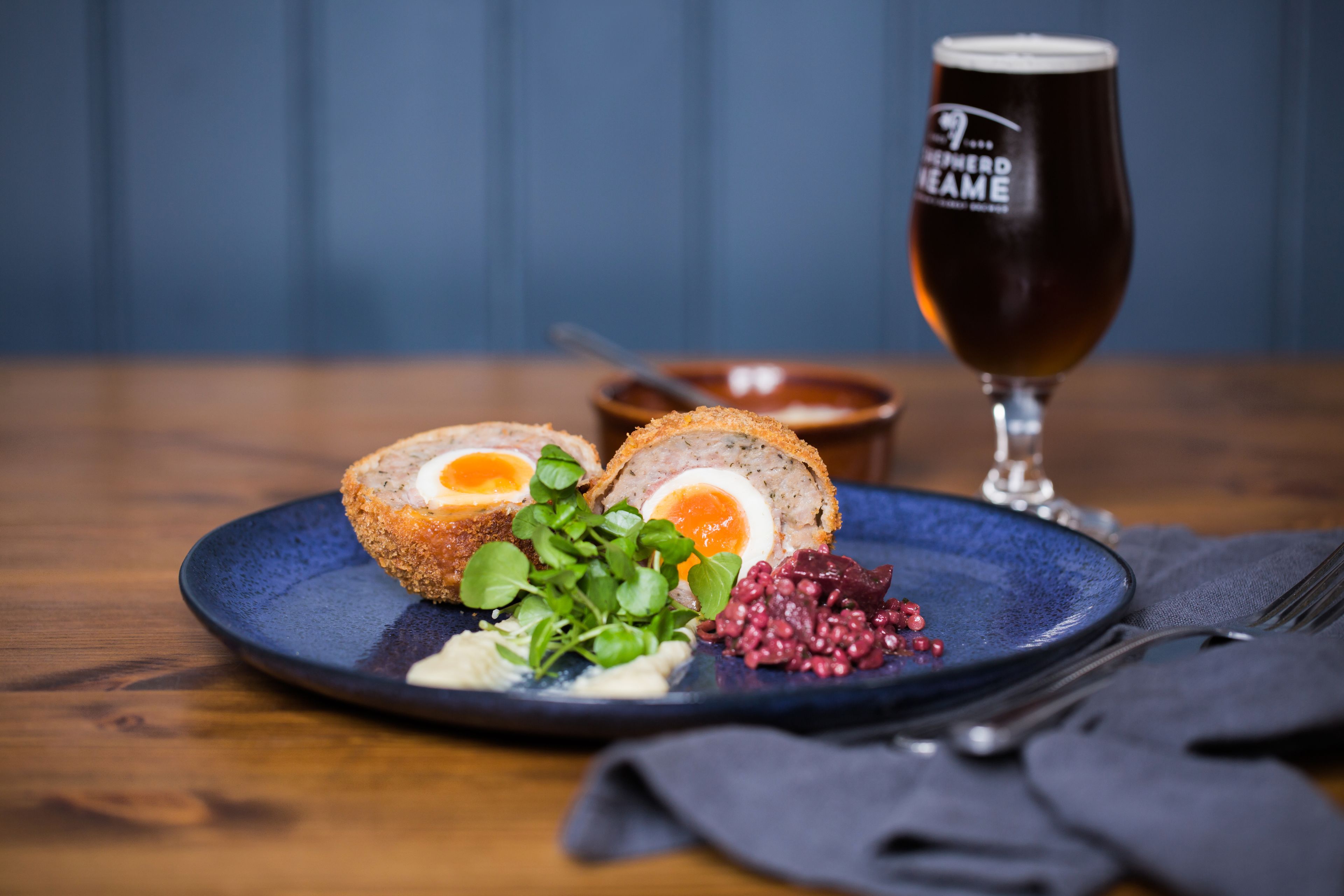 A plate with a Scotch egg, greens, and red berries, accompanied by a glass of beer on a wooden table.