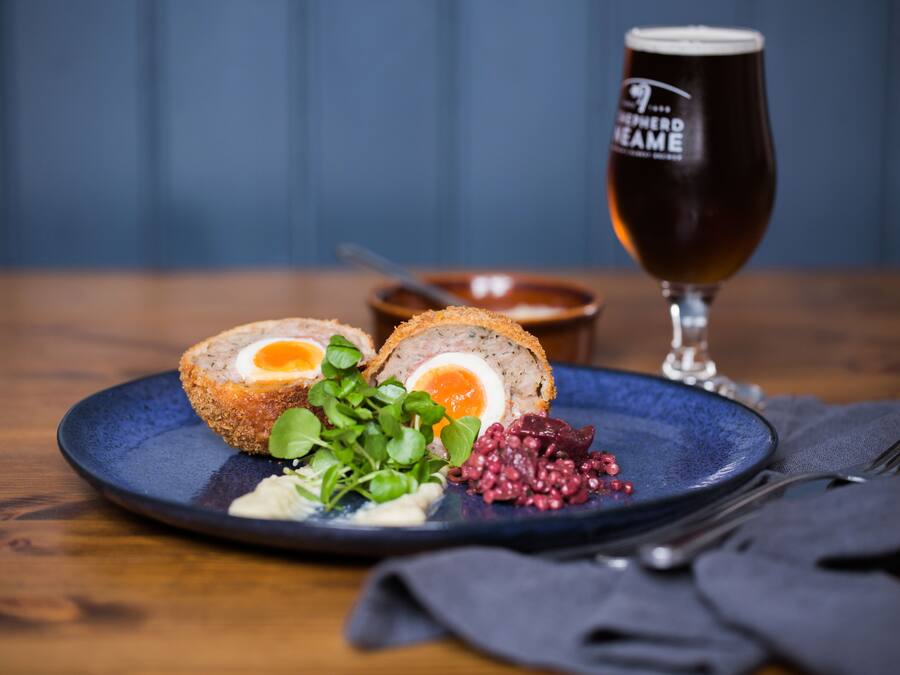 A plate with a Scotch egg, greens, and red berries, accompanied by a glass of beer on a wooden table.