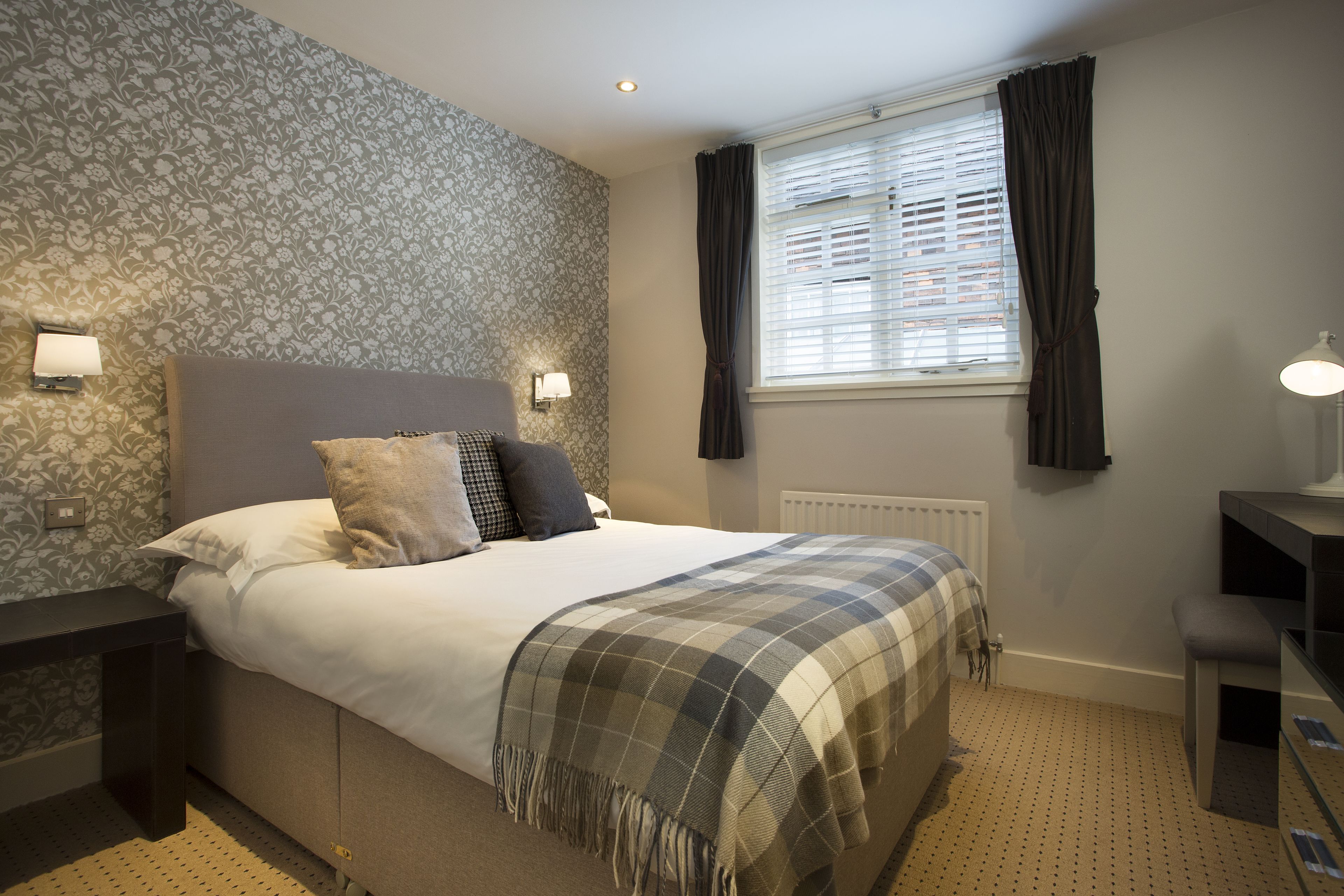 A bedroom with a bed, bedside lamps, a window with curtains, a radiator, and a desk with a lamp.